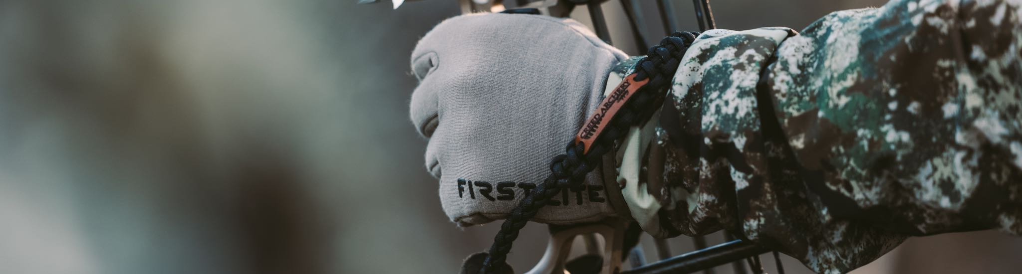 Close-up of gray "FIRST LITE" bow glove and black "CREED ARCHERY" paracord sling.