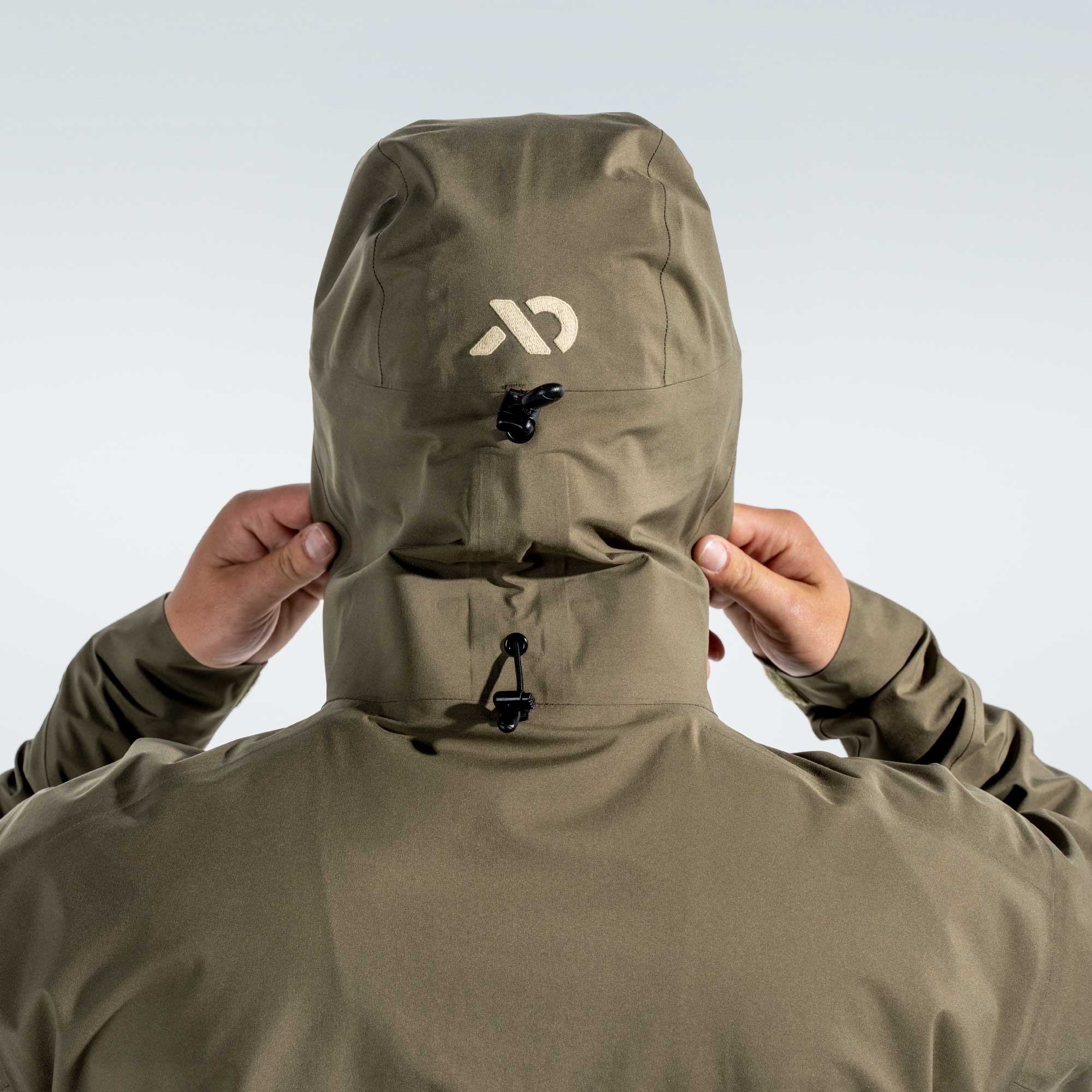 A person wearing an olive green hooded jacket, viewed from the back, adjusting the hood with both hands. A stylized white logo is printed on the back of the hood, and the jacket has black drawcord closures.