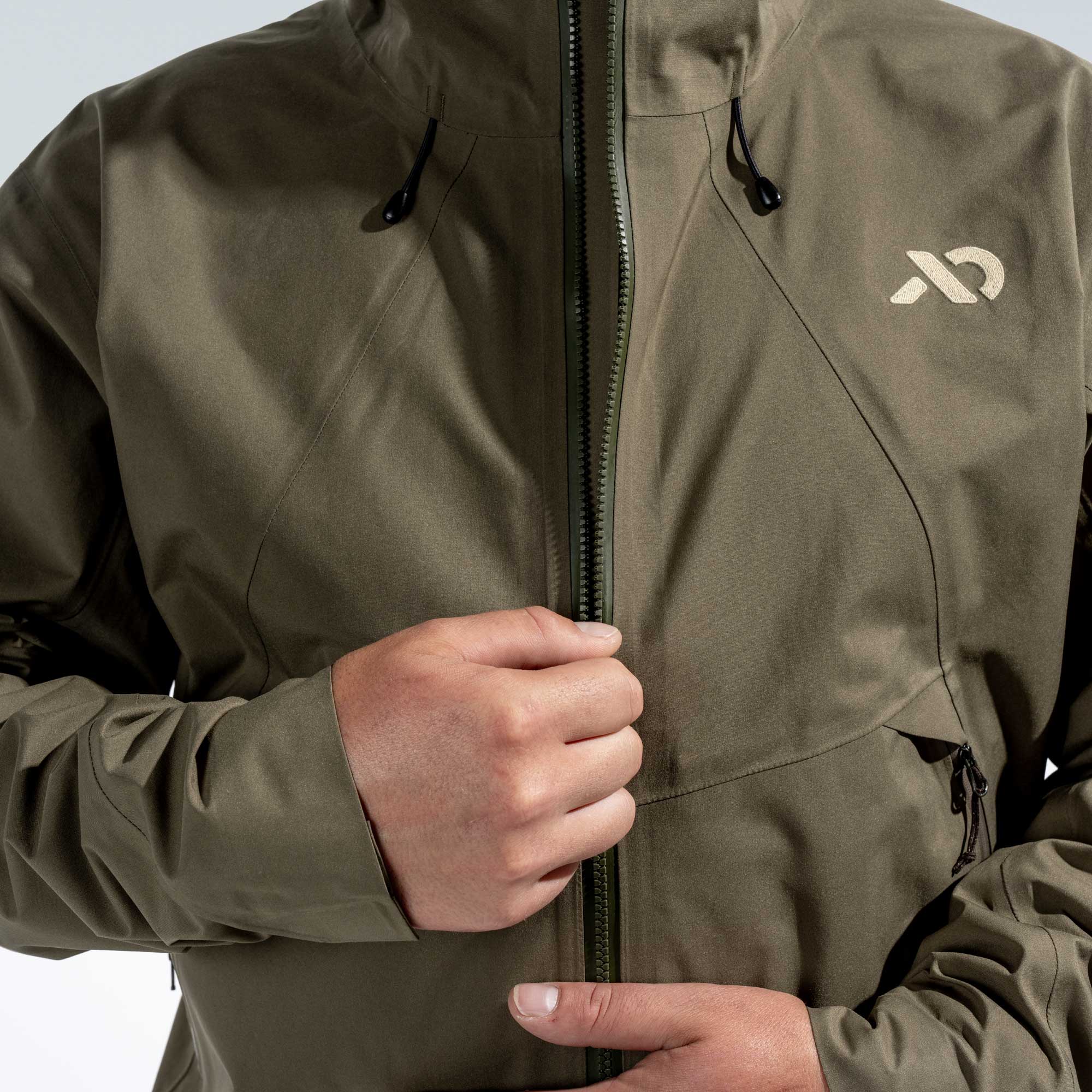 Close-up of a person wearing an olive green technical jacket, pulling the front zipper with both hands. A light beige logo is visible on the left chest.