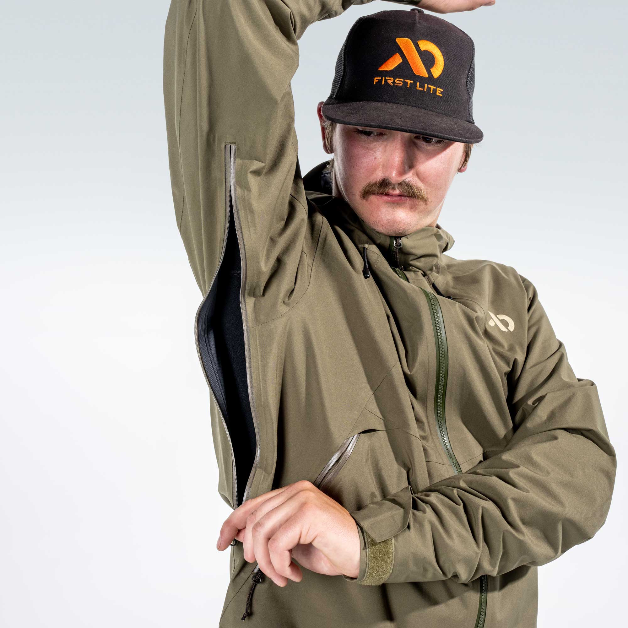 A man wearing an olive green waterproof jacket and a black First Lite cap, posing with one arm raised against a light gray background.
