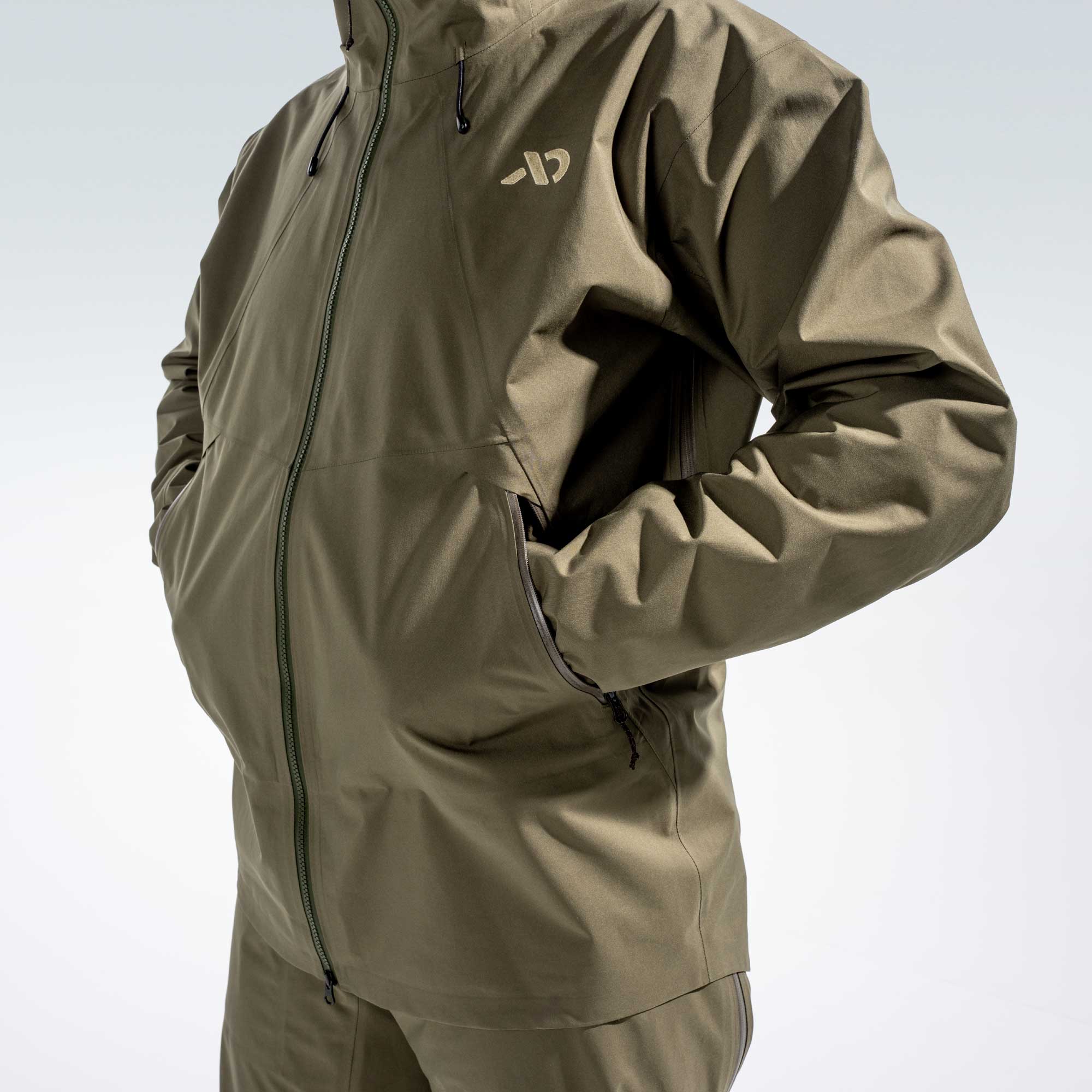 A close-up of a person wearing an olive green waterproof jacket with a front zipper, zippered side pocket, and a small white stylized logo on the chest, set against a light gray studio background.
