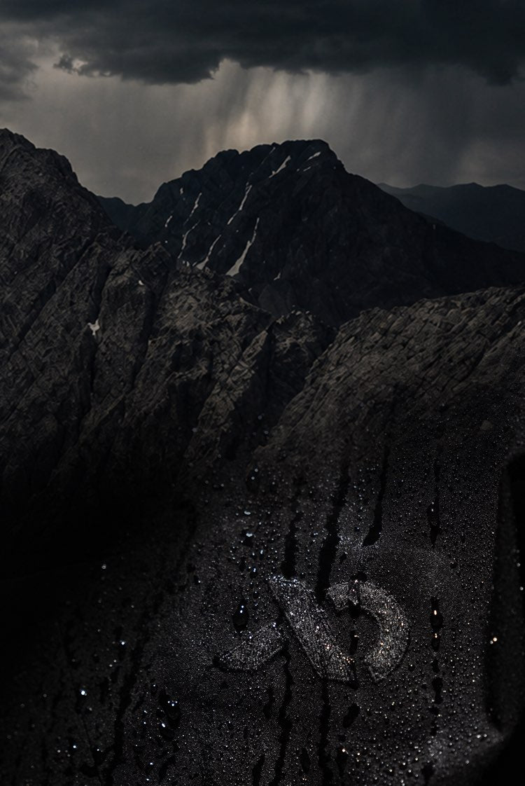Dark dramatic mountain landscape with storm clouds, rain shafts, and a winding road or path cutting through the rocky terrain.