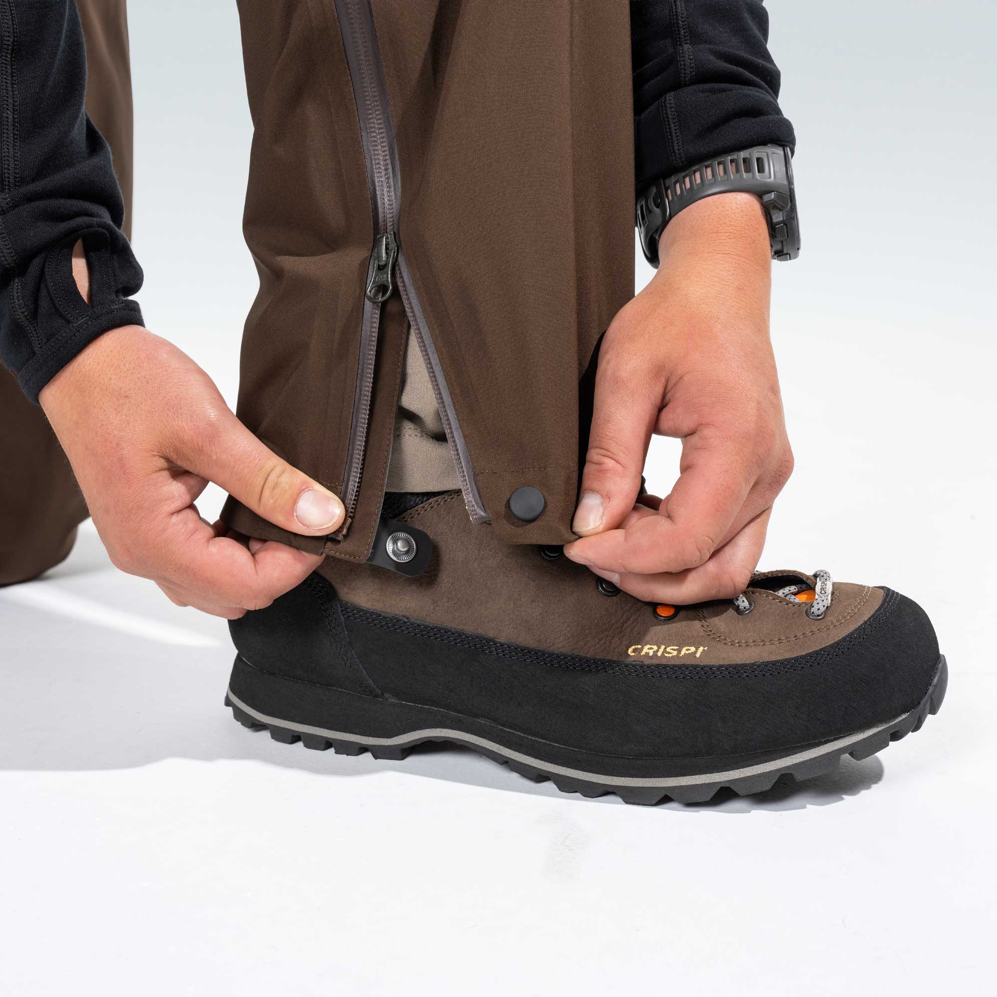 A close-up of a person's hands fastening a brown and black Crispi hiking boot with a quick-release pant snap on brown pants, set against a clean white studio background.