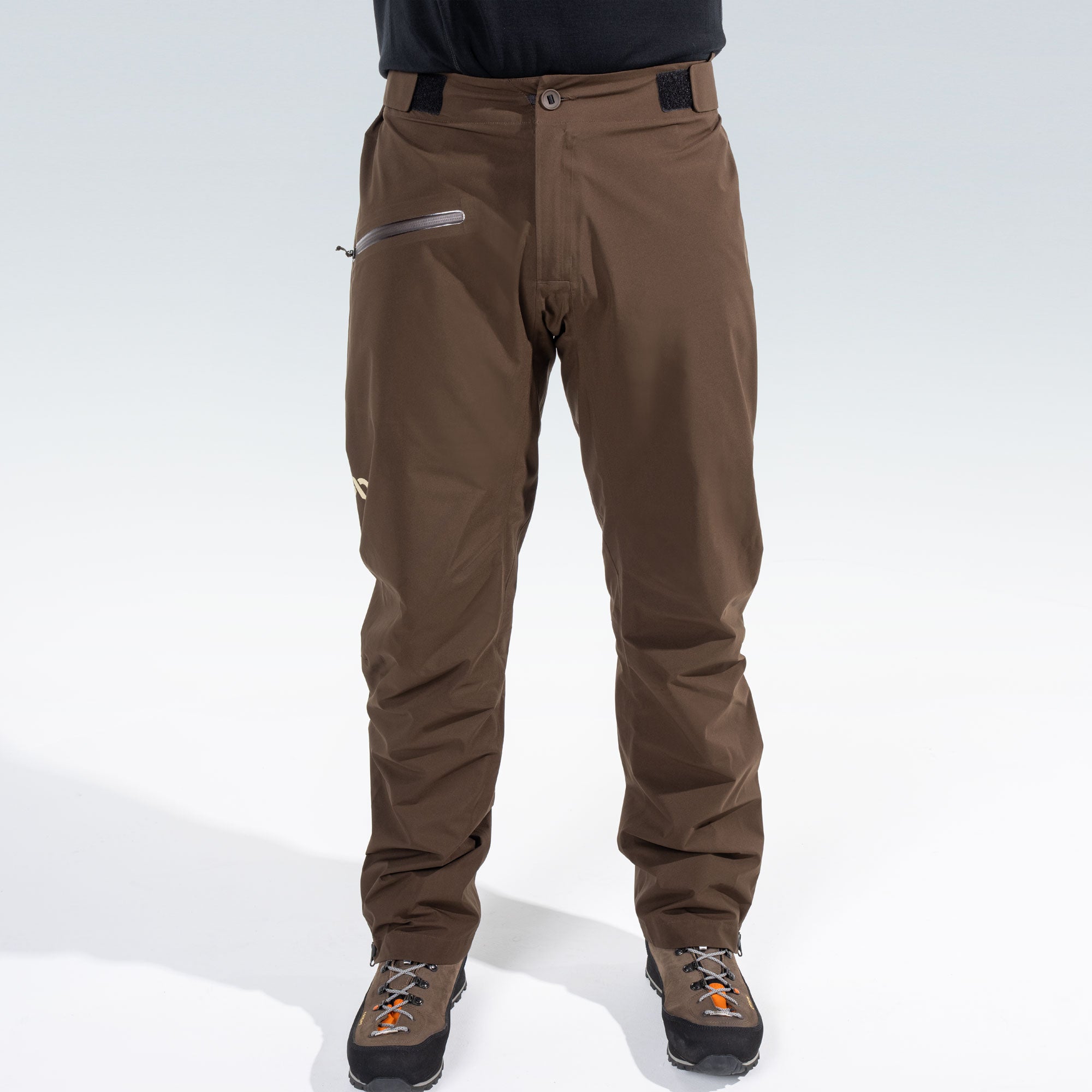 A person from the waist down wearing brown technical rain pants with a zippered pocket and brown hunting boots, shown against a light gray studio background.