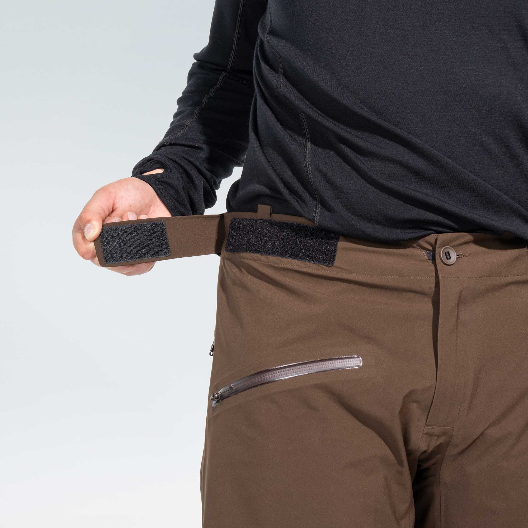 A close-up of a person wearing brown technical pants and a black long-sleeve top, demonstrating a brown elastic belt with hook-and-loop fastening around the waistband of the pants.