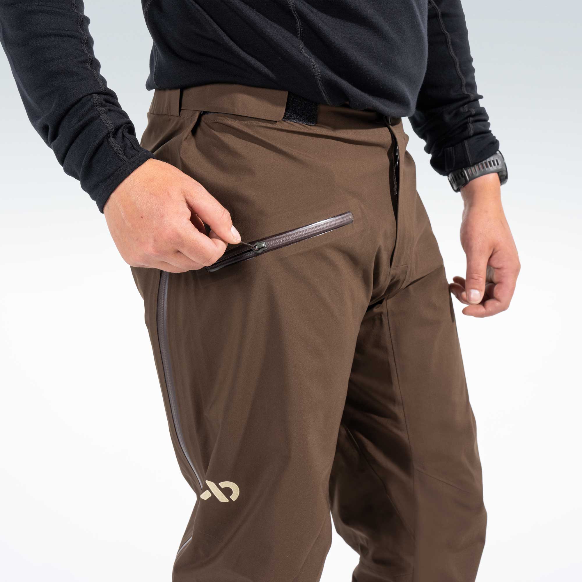 A close-up of a person wearing brown rain pants with a black long-sleeve top, demonstrating a zippered pocket on the thigh. The brand logo on the lower leg is visible.