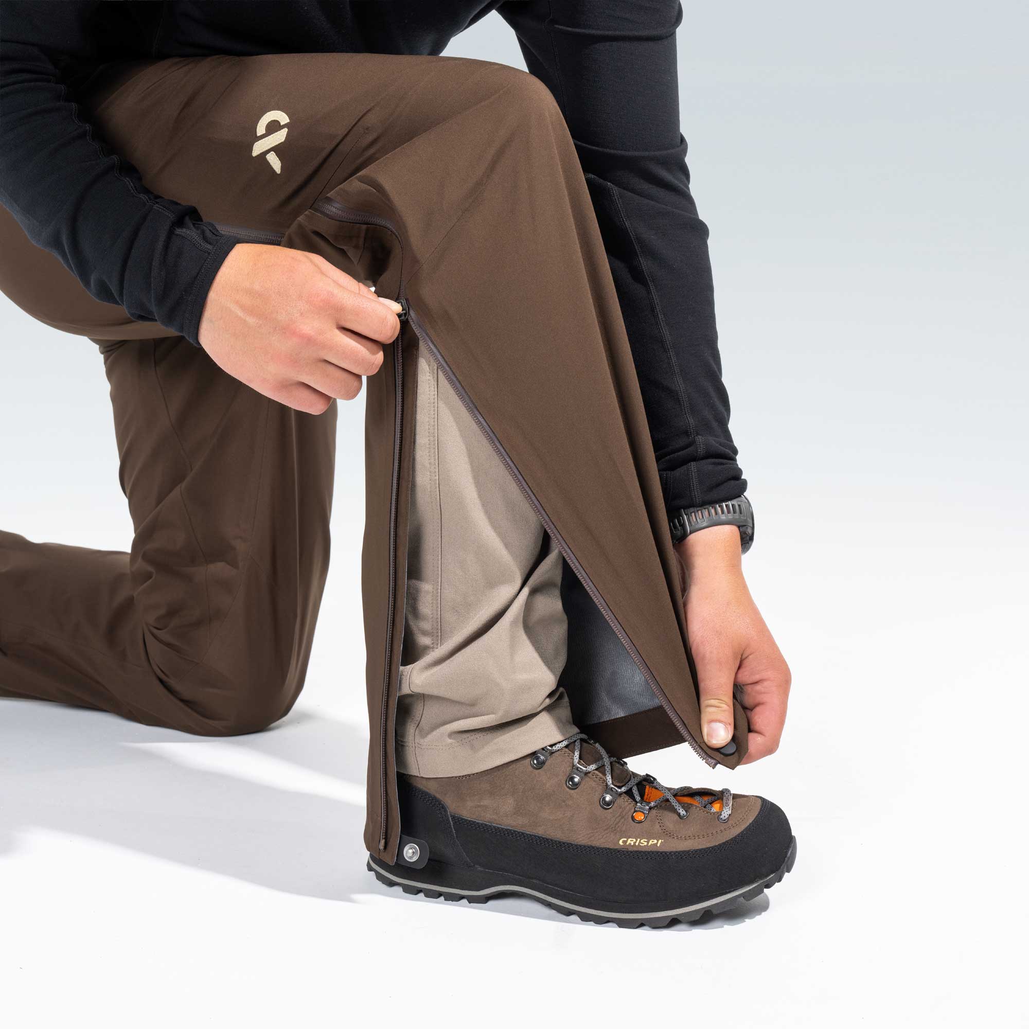 A close-up of a person kneeling and pulling down a brown waterproof pants liner over their boot to demonstrate a waterproofing feature. The person is wearing brown pants, a black long-sleeve shirt, and a brown and black Crispi hiking boot.