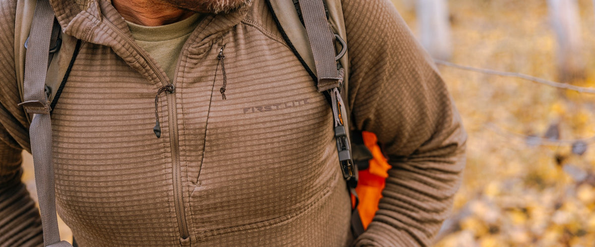 A close-up of a person wearing a brown waffle-knit fleece zip-up jacket with a visible First Lite logo on the chest, along with a backpack in an outdoor autumn setting.