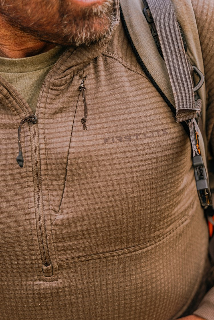 A close-up of a person wearing a brown waffle-knit fleece zip-up jacket with a visible First Lite logo on the chest, along with a backpack in an outdoor autumn setting.
