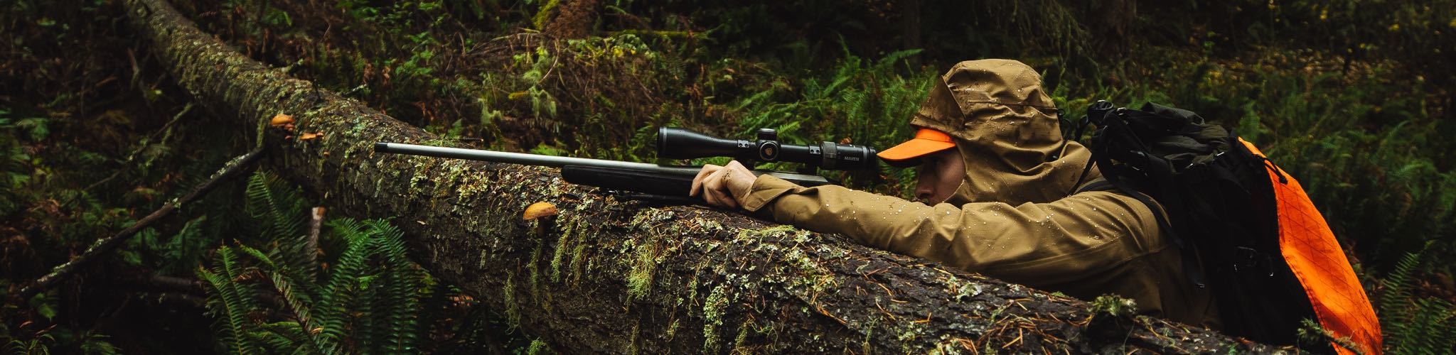 Man in rain jacket looks down gun resting on log