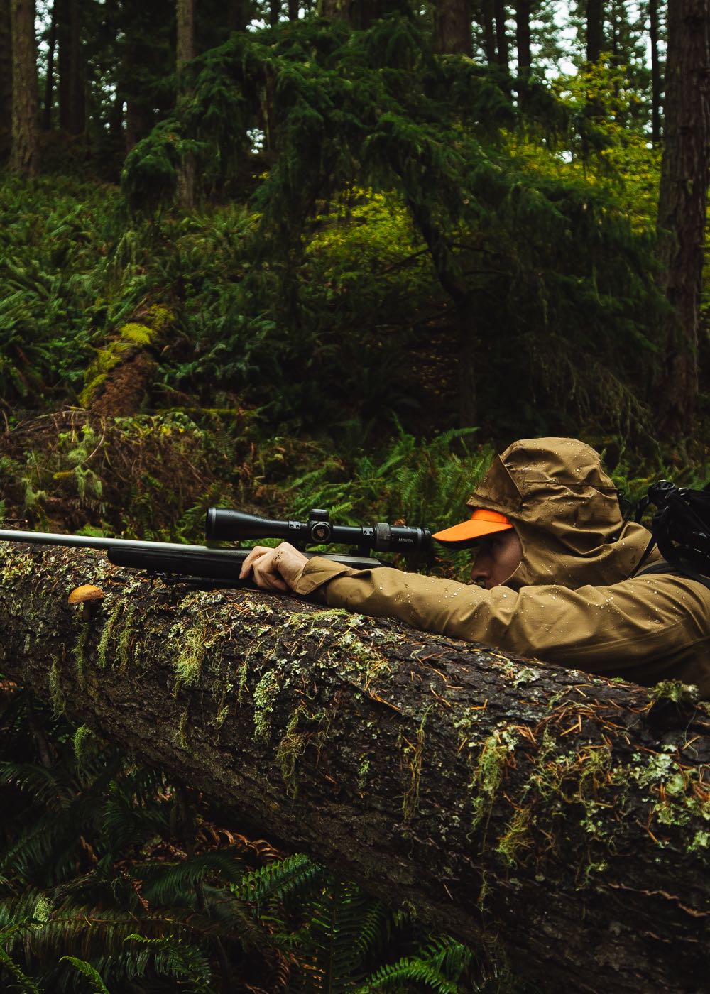 Man in rain jacket looks down gun resting on log