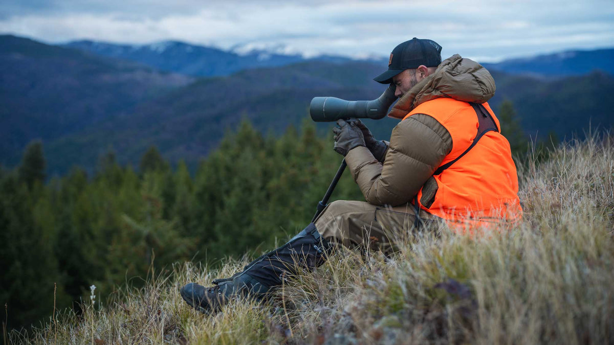 Hunter in a bright orange vest using a spotting scope to glass rolling, timbered mountains. 