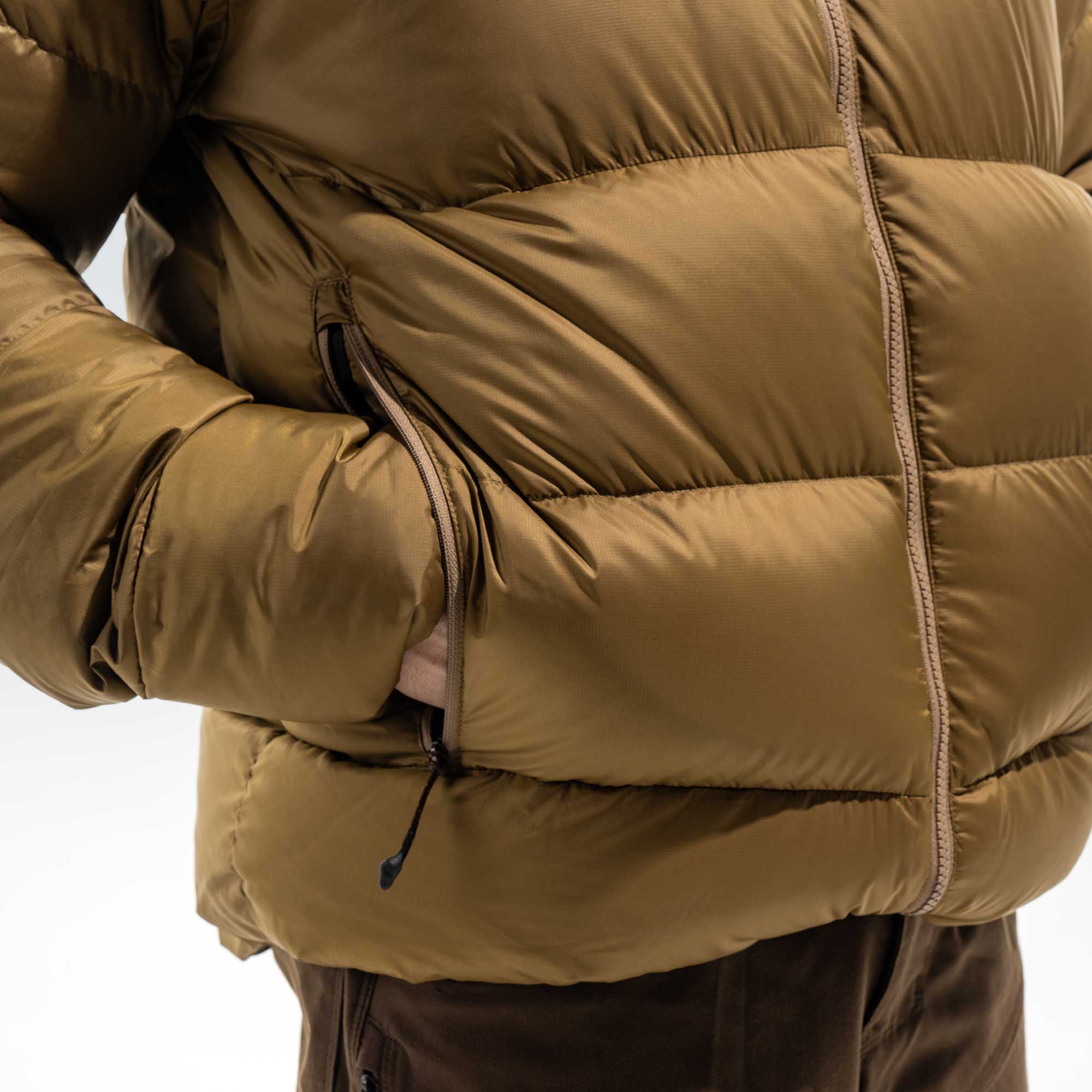 Close-up of a person wearing a padded quilted jacket in a camel brown color, showing the chest, zipper, and side pocket with a hand inside.