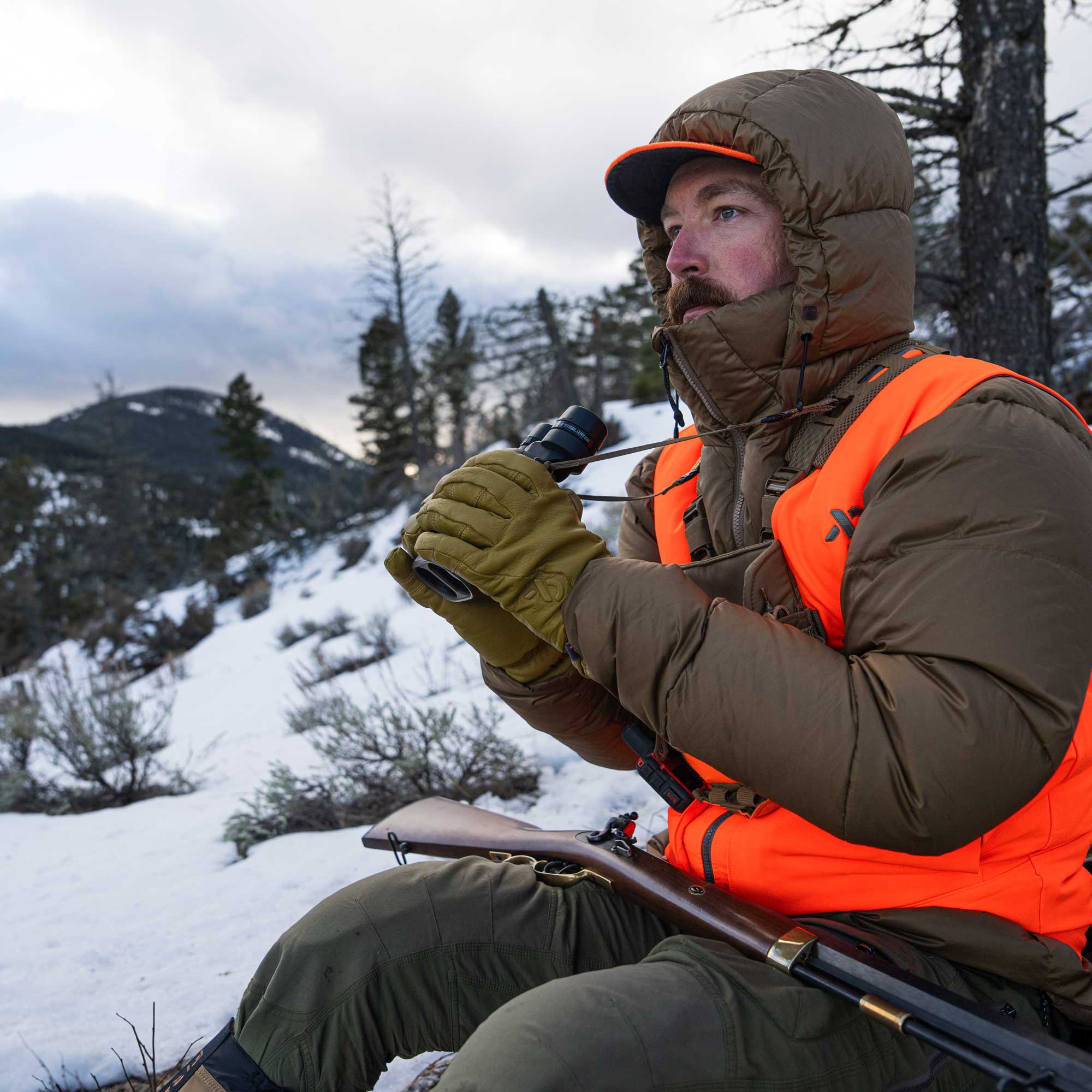 A man in a snowy outdoor setting wearing a brown puffer jacket, bright orange hunting vest, olive green gloves, and holding a shotgun.