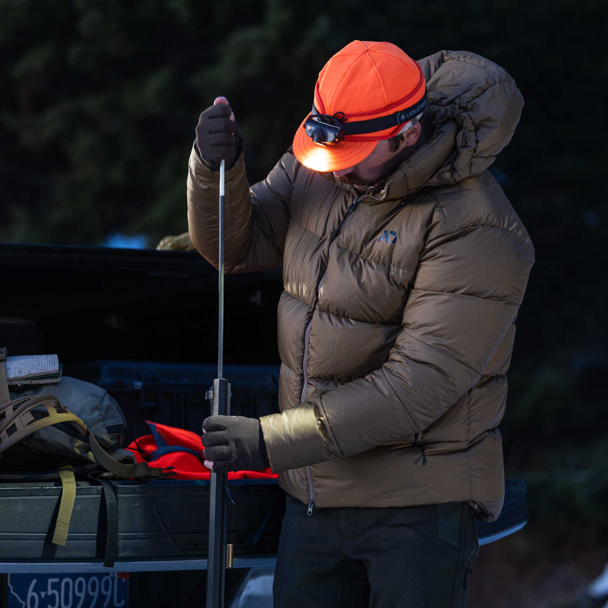 A person wearing a tan puffer jacket, orange cap, and headlamp stands by a truck at night, holding a muzzleloader. The jacket has a small visible First Lite logo on the chest.