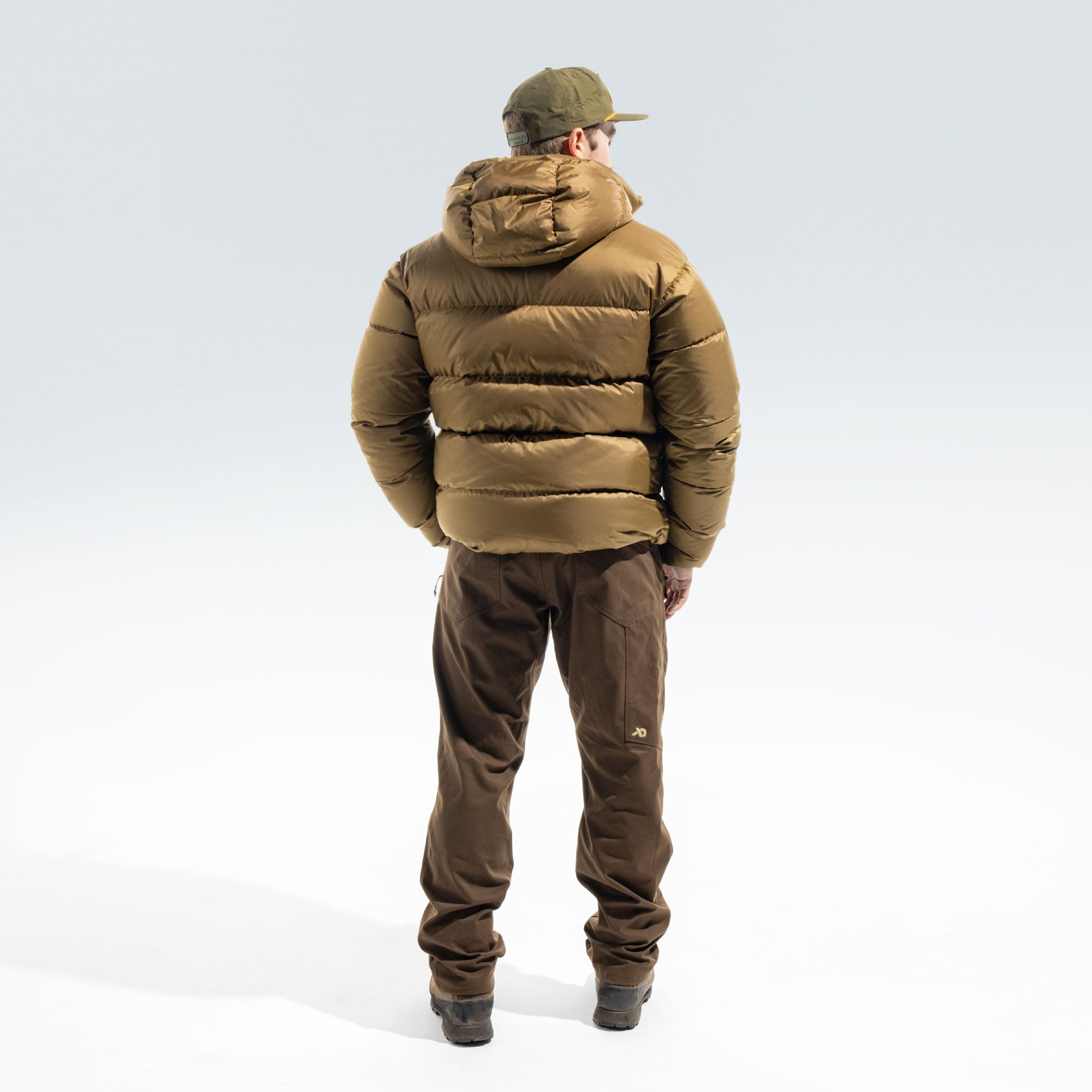 A man viewed from behind wearing a brown puffer jacket, brown pants, and an olive green cap, standing against a light gray studio background.
