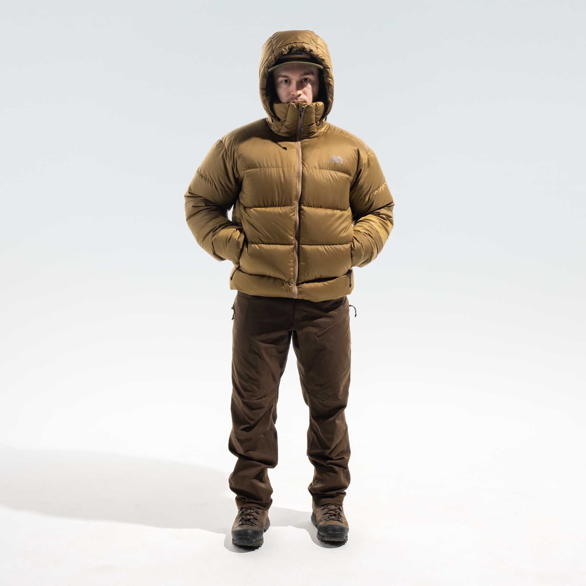 A man wearing a tan hooded puffer jacket, brown pants, and dark boots, standing against a light gray studio background.