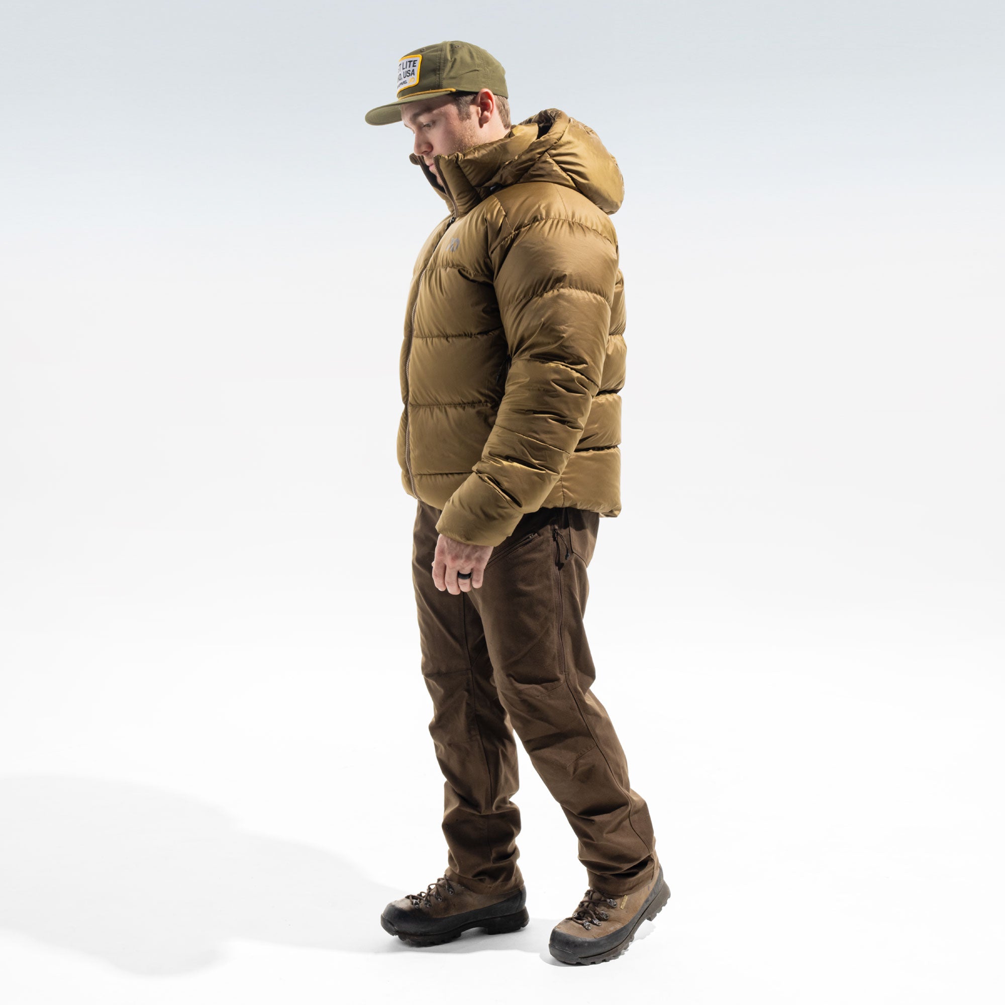 A man wearing a tan hooded puffer jacket, brown pants, brown hiking boots, and an olive green cap, standing in profile against a light gray studio background.