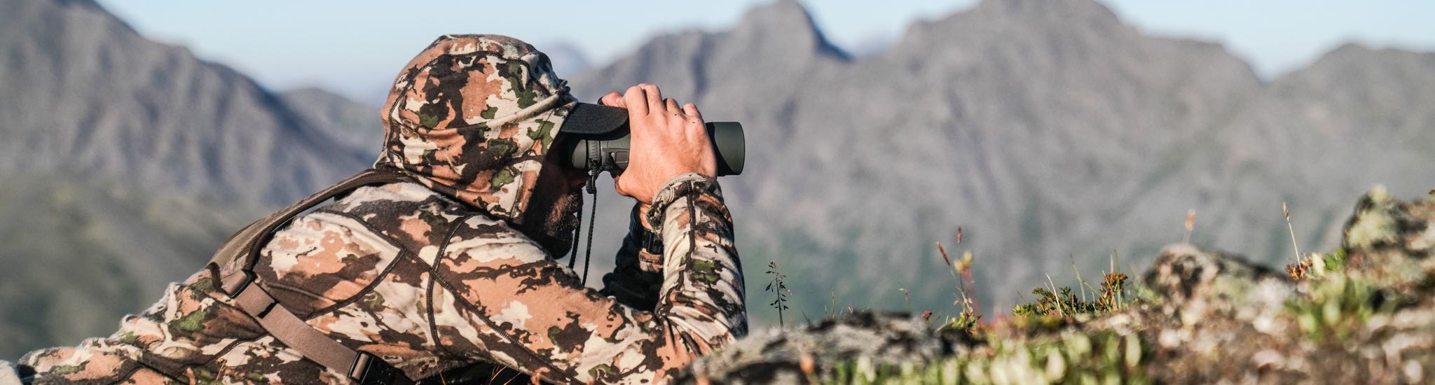 Hunter in hooded camouflage top observing mountain landscape through binoculars.