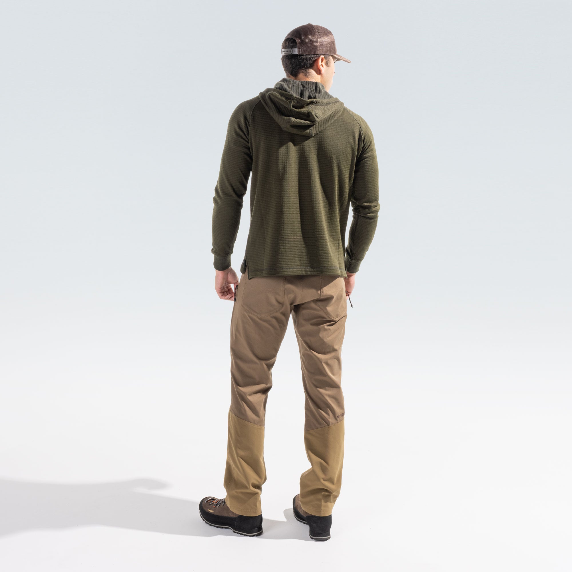 Man wearing tan brush pants and a green grid hoody, standing facing away from the camera on a white background.