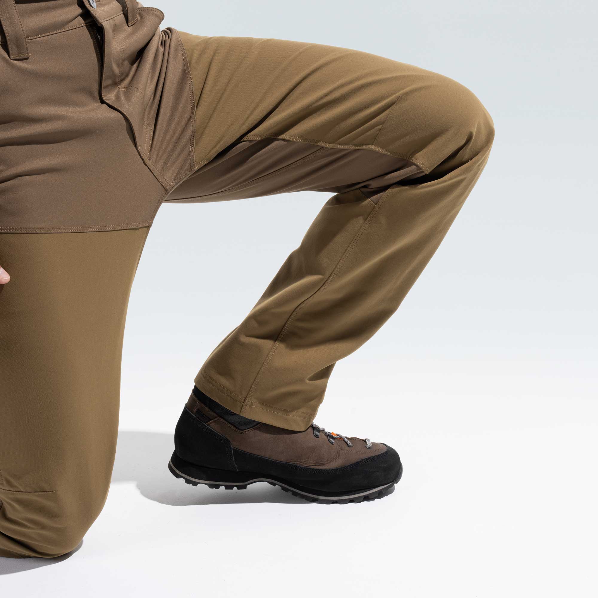 A close-up of a person wearing brown and black hiking boots with tan pants, shown from the waist down against a clean white studio background.