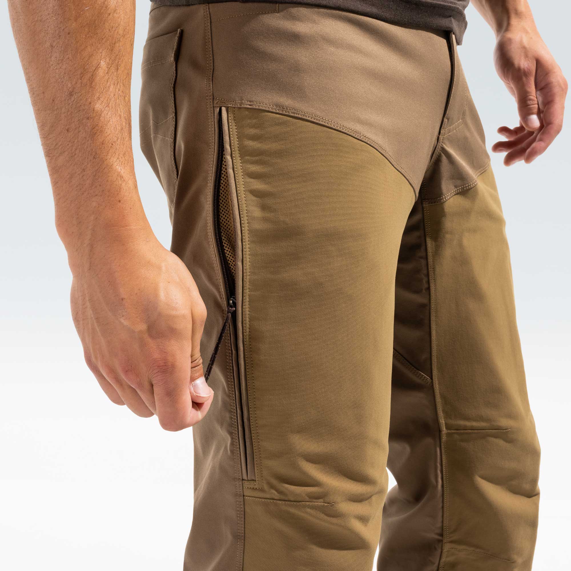 Close-up of a person wearing tan technical outdoor pants, showing a zippered pocket on the thigh and the side panel construction against a light studio background.
