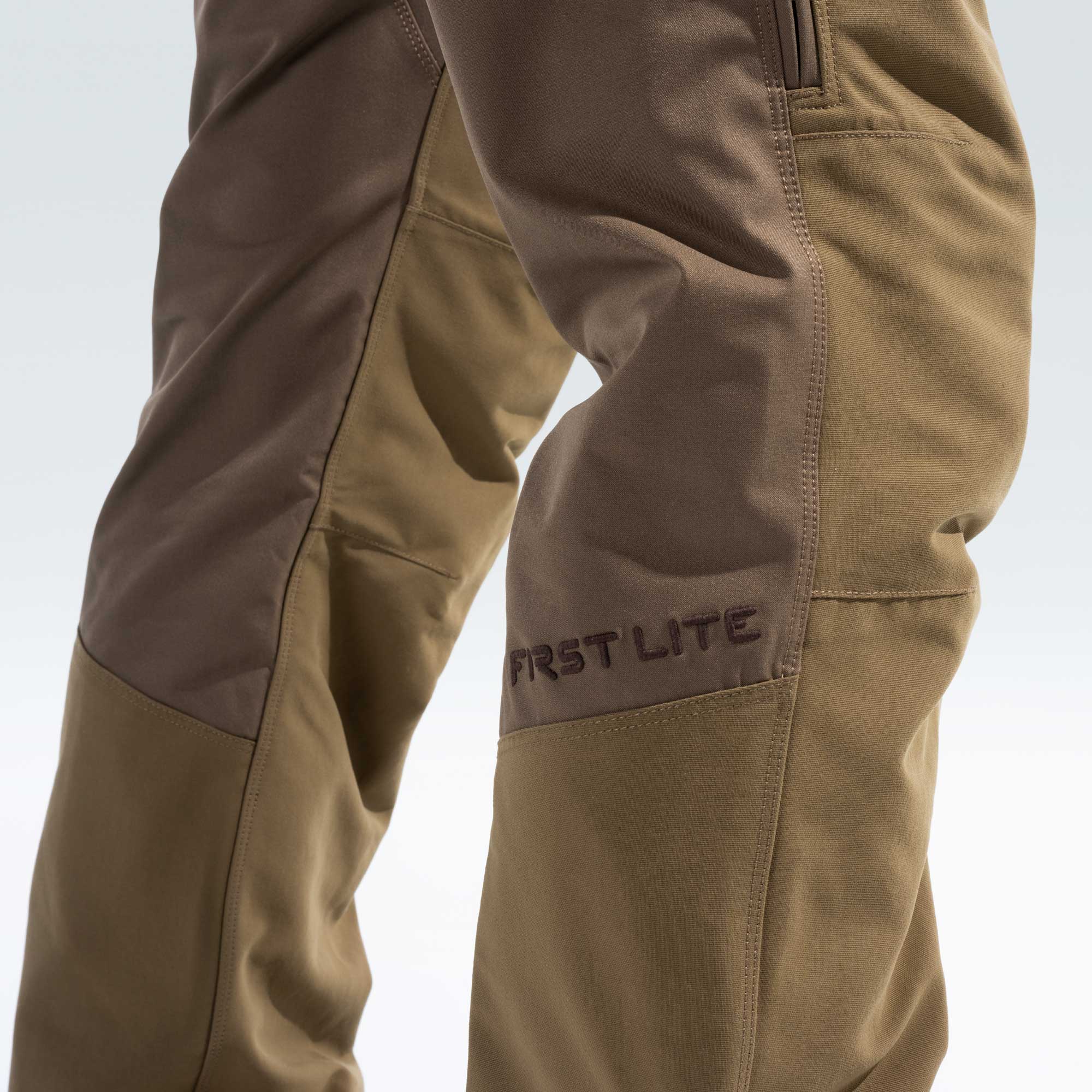 Close-up of a person's lower legs wearing First Lite outdoor pants in a brown and khaki color-blocked design, with the brand name 'FIRST LITE' embroidered on the side.