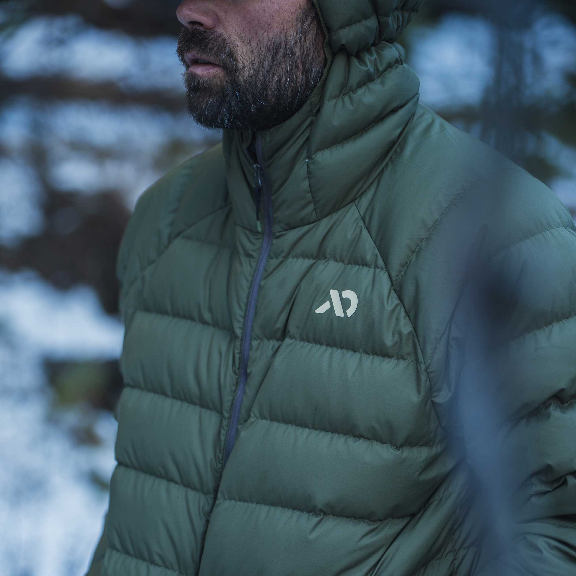 A close-up of a man wearing an olive green hooded puffer jacket outdoors, with snow and blurred trees in the background. A small white logo is visible on the left chest.