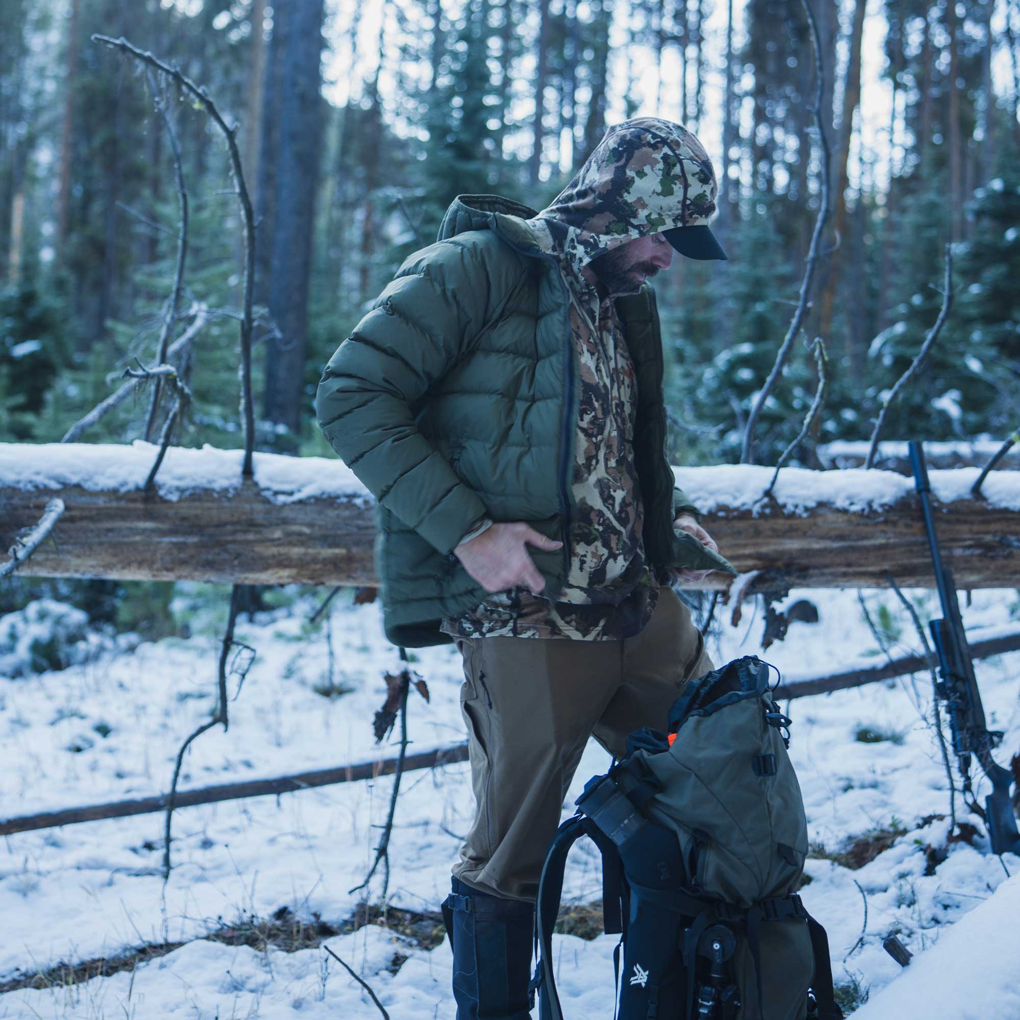 A man in a snowy forest wearing an olive green puffer jacket, camouflage hood, and brown pants, standing next to a large green and black backpack.