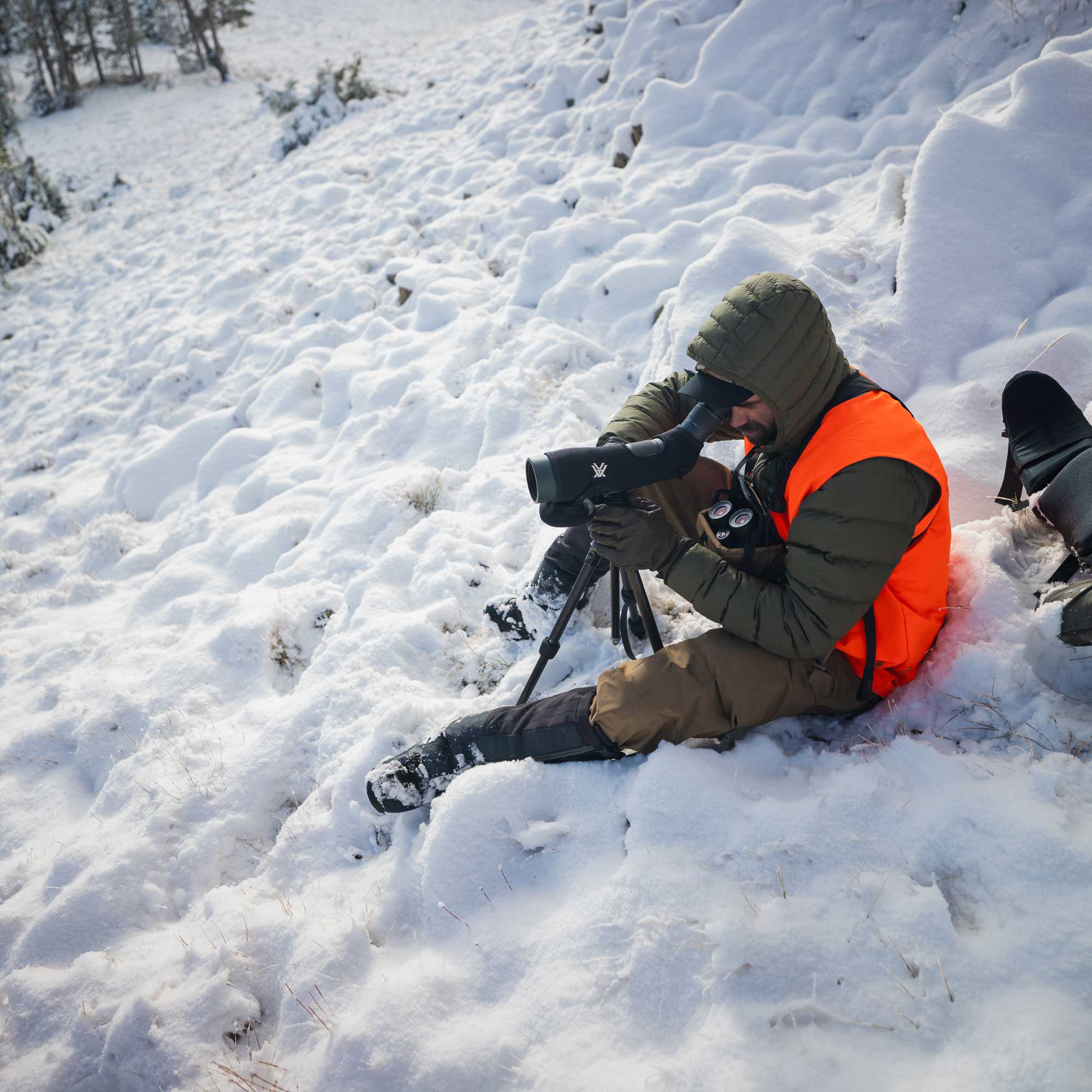 A person in winter clothing sits in deep snow, using a spotting scope mounted on a tripod in an outdoor snowy landscape.