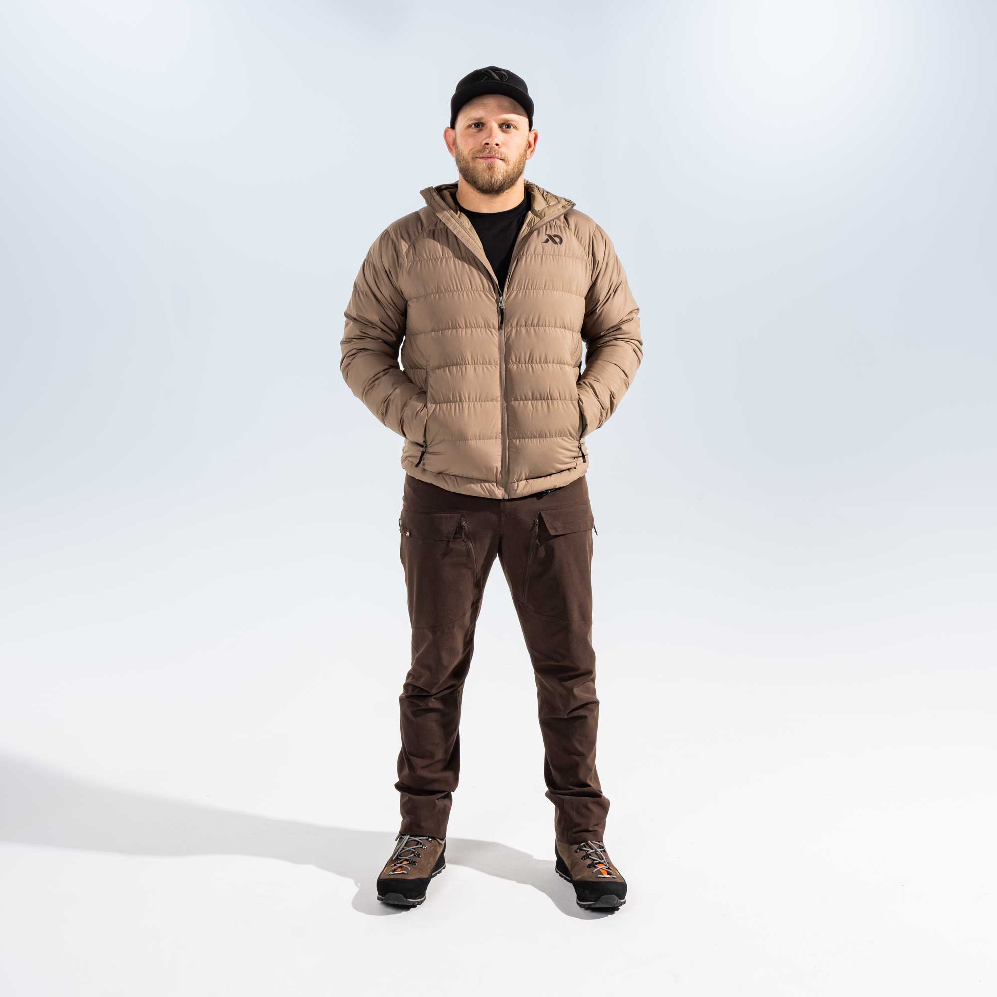 A man wearing a light brown puffer jacket, dark brown pants, and a black cap, standing against a light gray studio background.