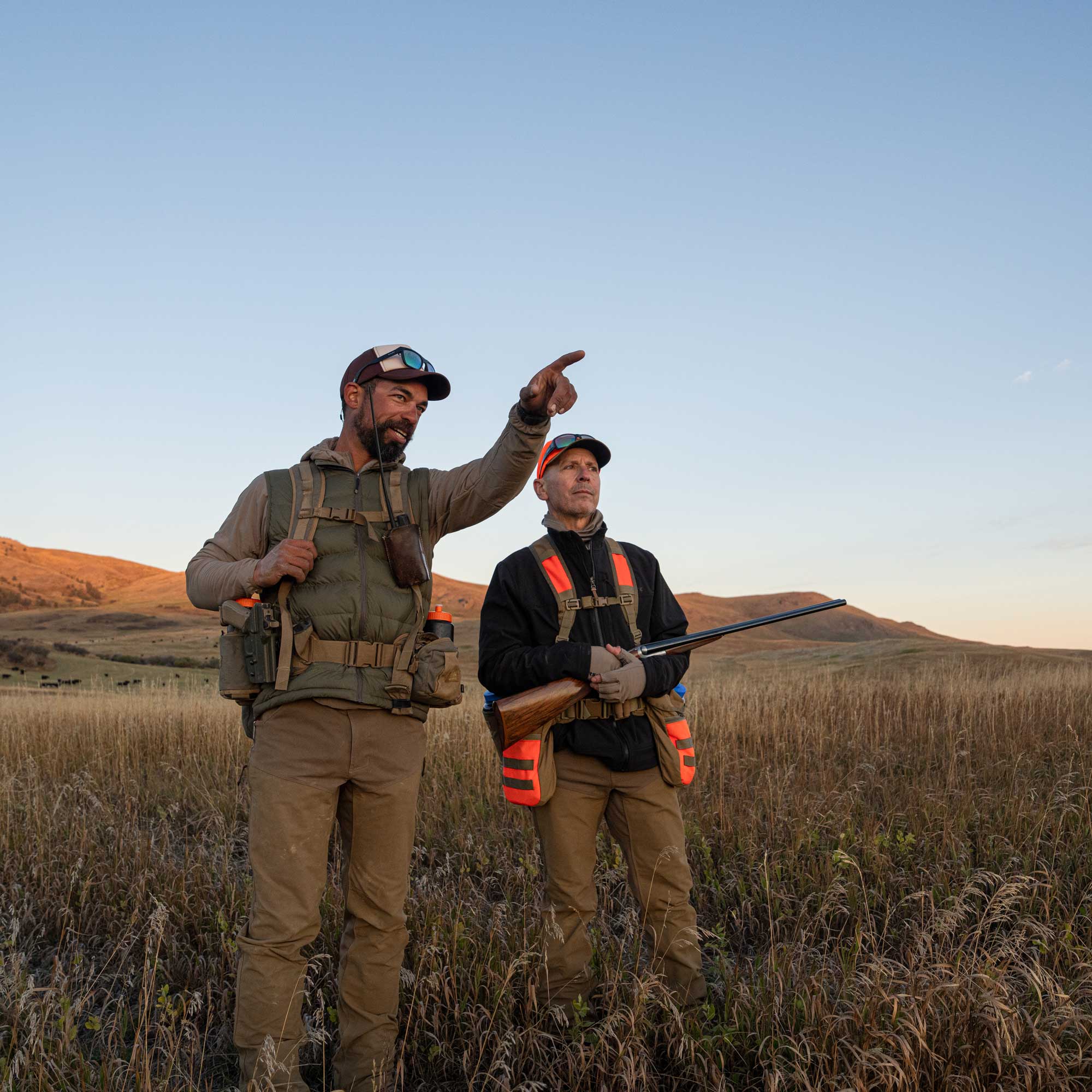 Two men wearing upland vests and on a bird hunt, standing on the prairie wearing upland hunting clothing. 