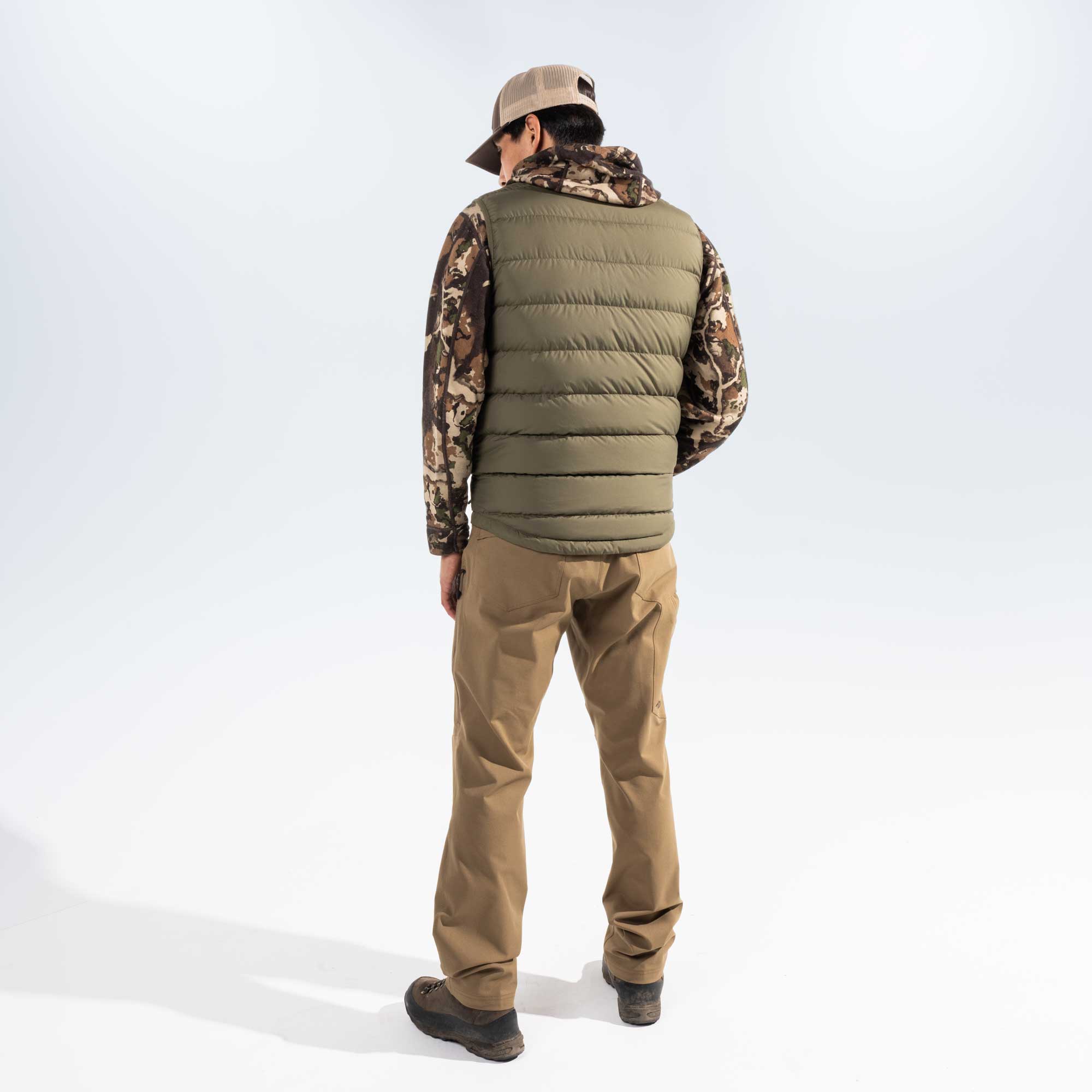 A man is shown from behind wearing a sage green quilted puffer vest over a camouflage hoodie, tan pants, and a beige baseball cap, standing against a white studio background.