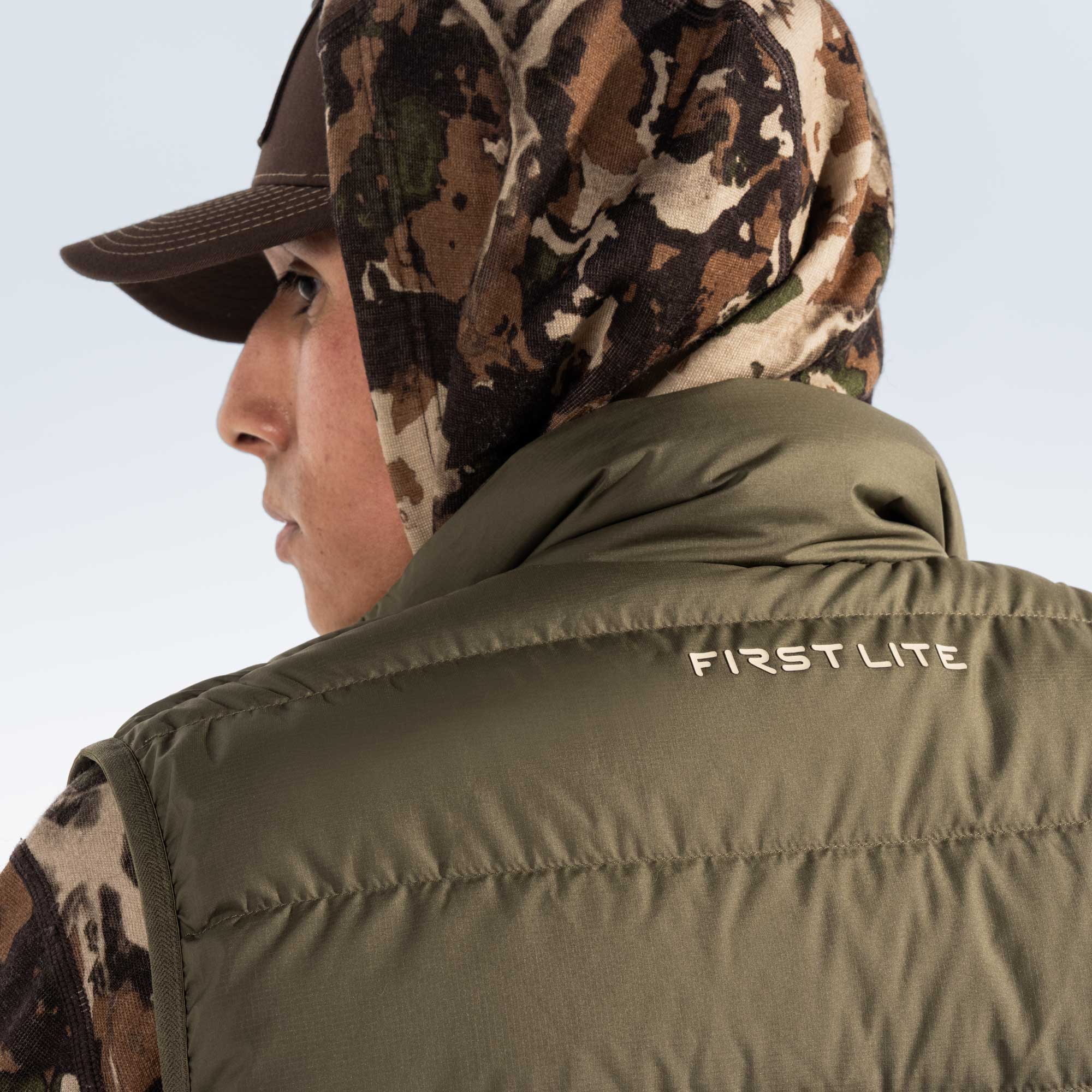 Reverse shot of a man with a hooded camouflage sweatshirt and olive green puffer vest, close up on the vest collar with text "First Lite". 