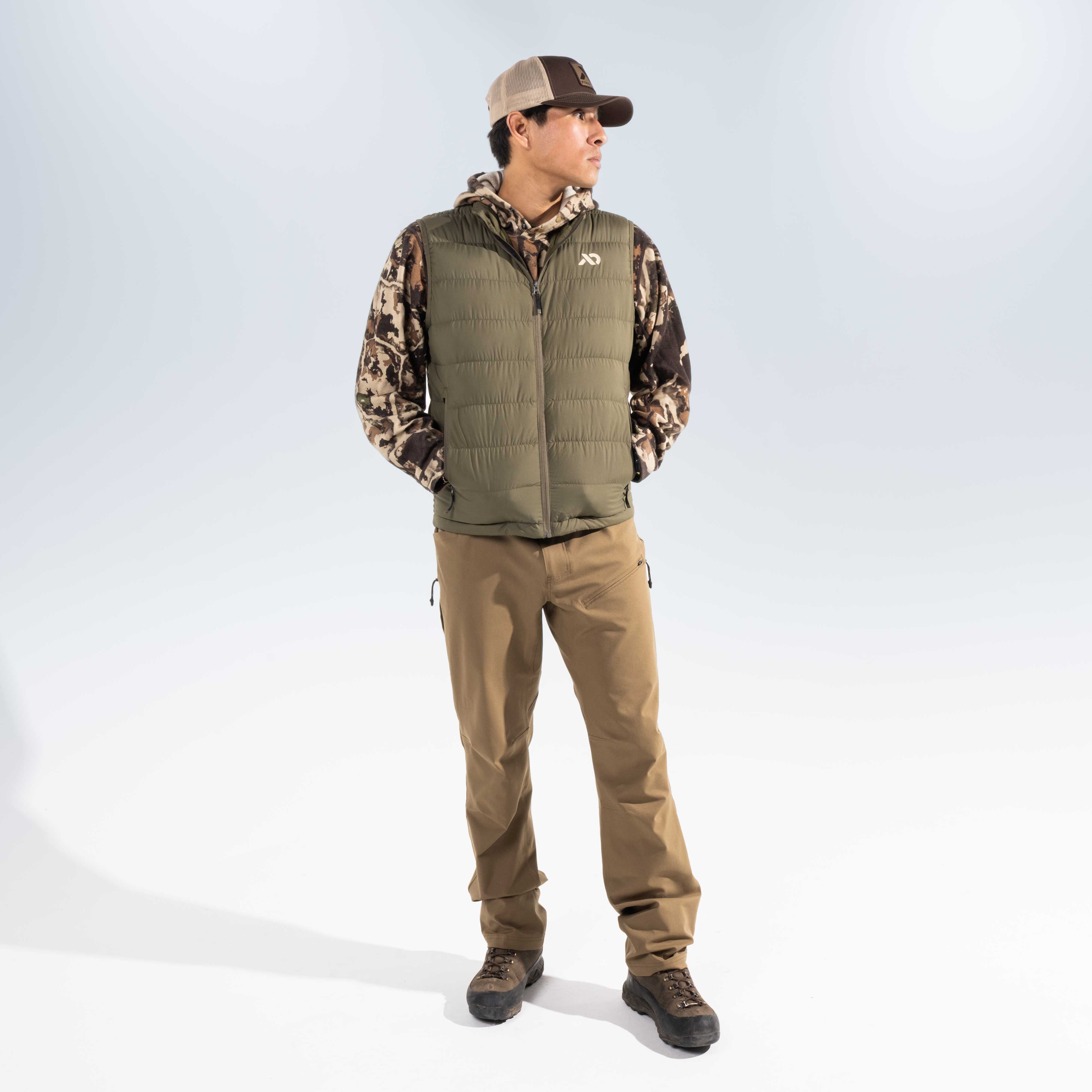 A man wearing a camouflage long-sleeve shirt under a quilted olive green vest, tan pants, and a baseball cap, standing against a light gray studio background.
