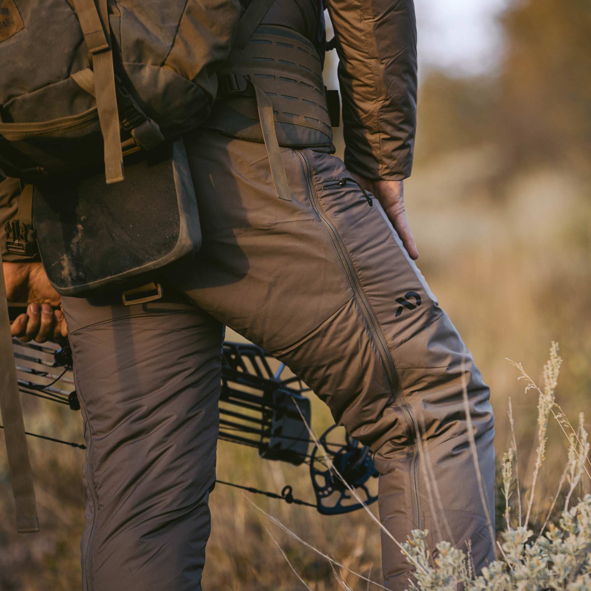 Man with a compound bow standing in sage brush with full length side zippers on a tan brown, insulated pant.
