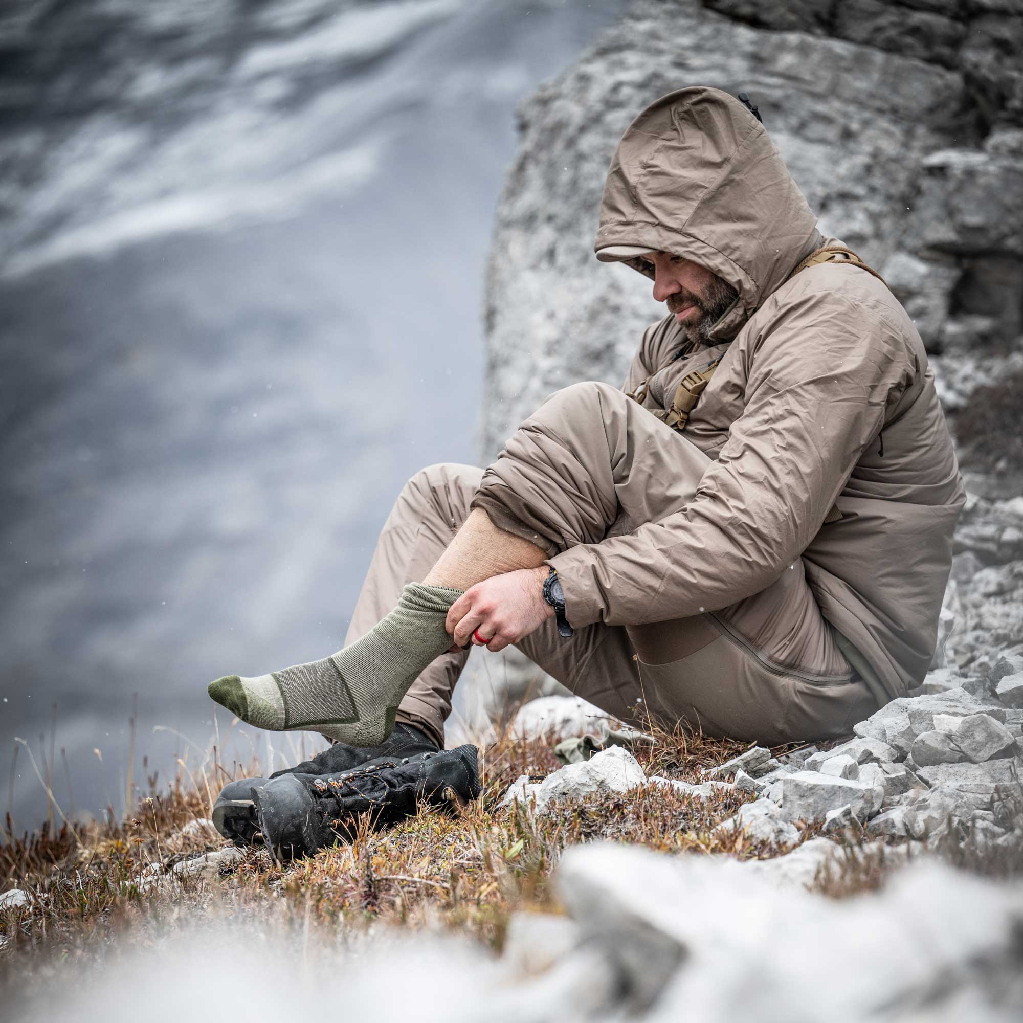 Man putting on a pair of socks in a rocky steppe with snow falling. 