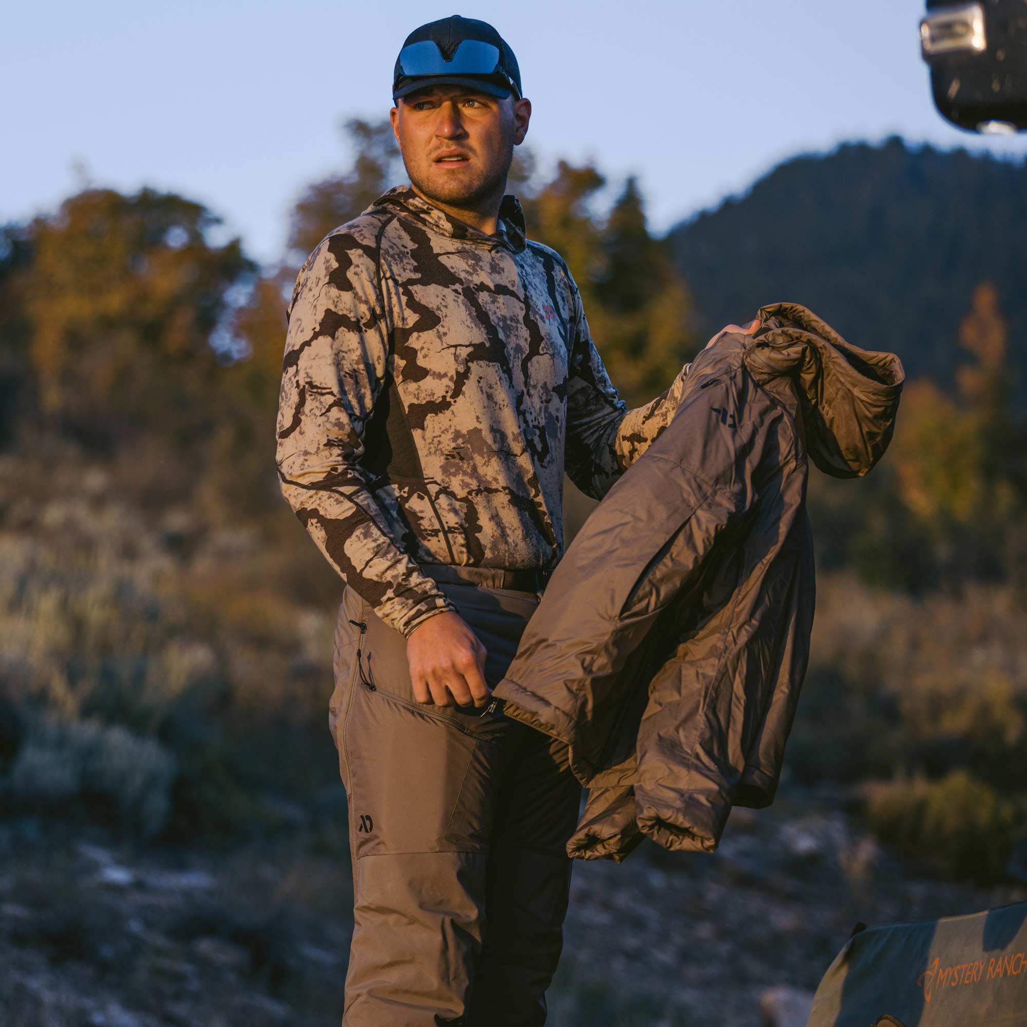 Man putting on a gray tan jacket and insulated pant as the sun rises. 