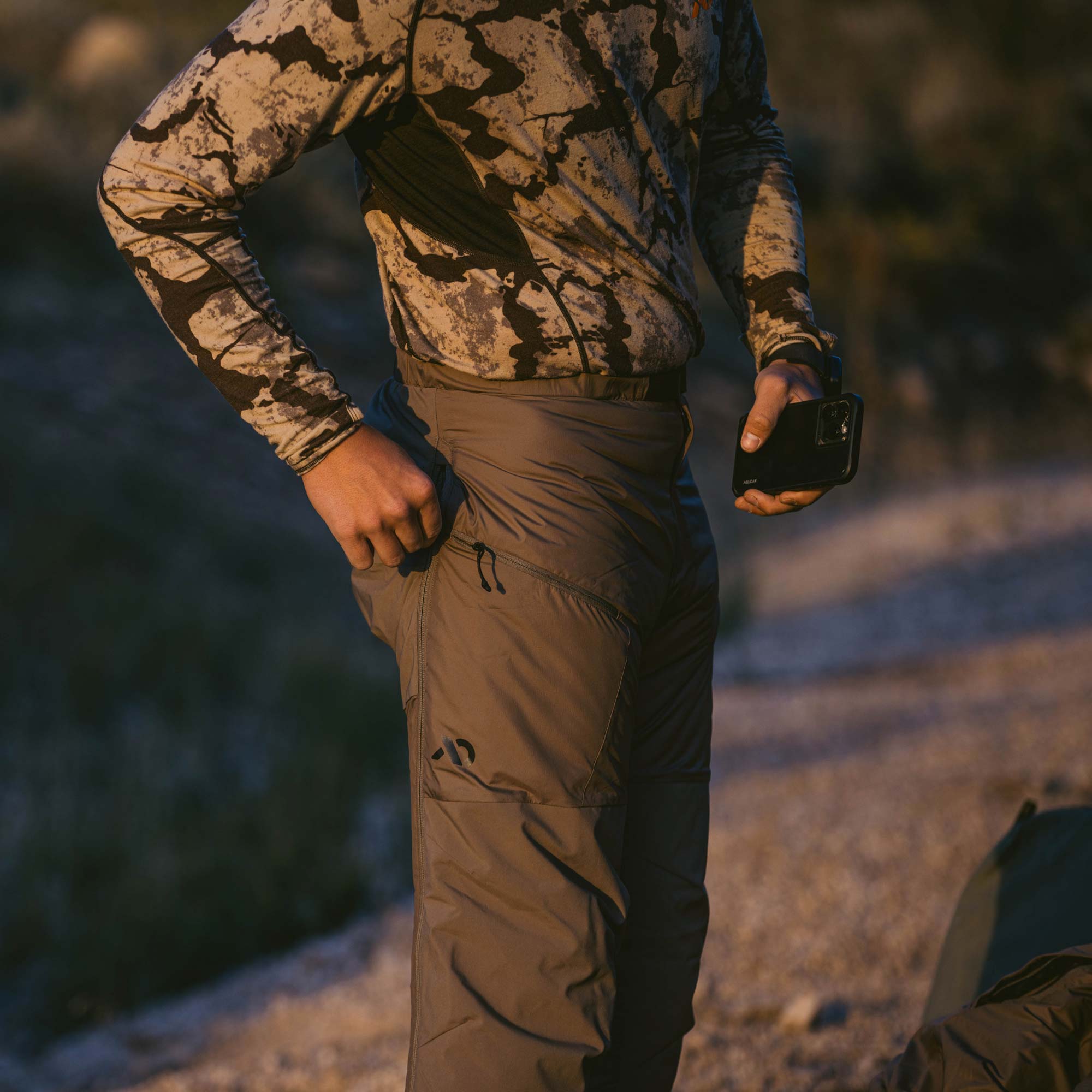 Man in camo putting his cell phone into the thigh pocket of a gray tan insulated puffy pant during early morning. 