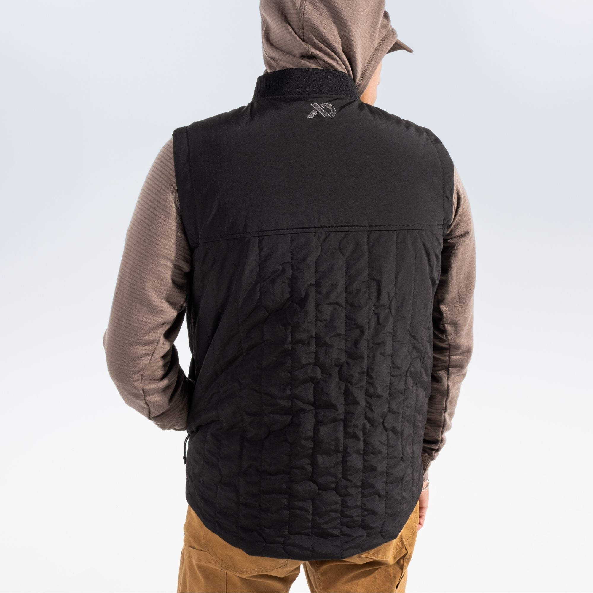 Man shown from behind wearing a black vest over a brown hoodie and tan pants standing against a white studio background.