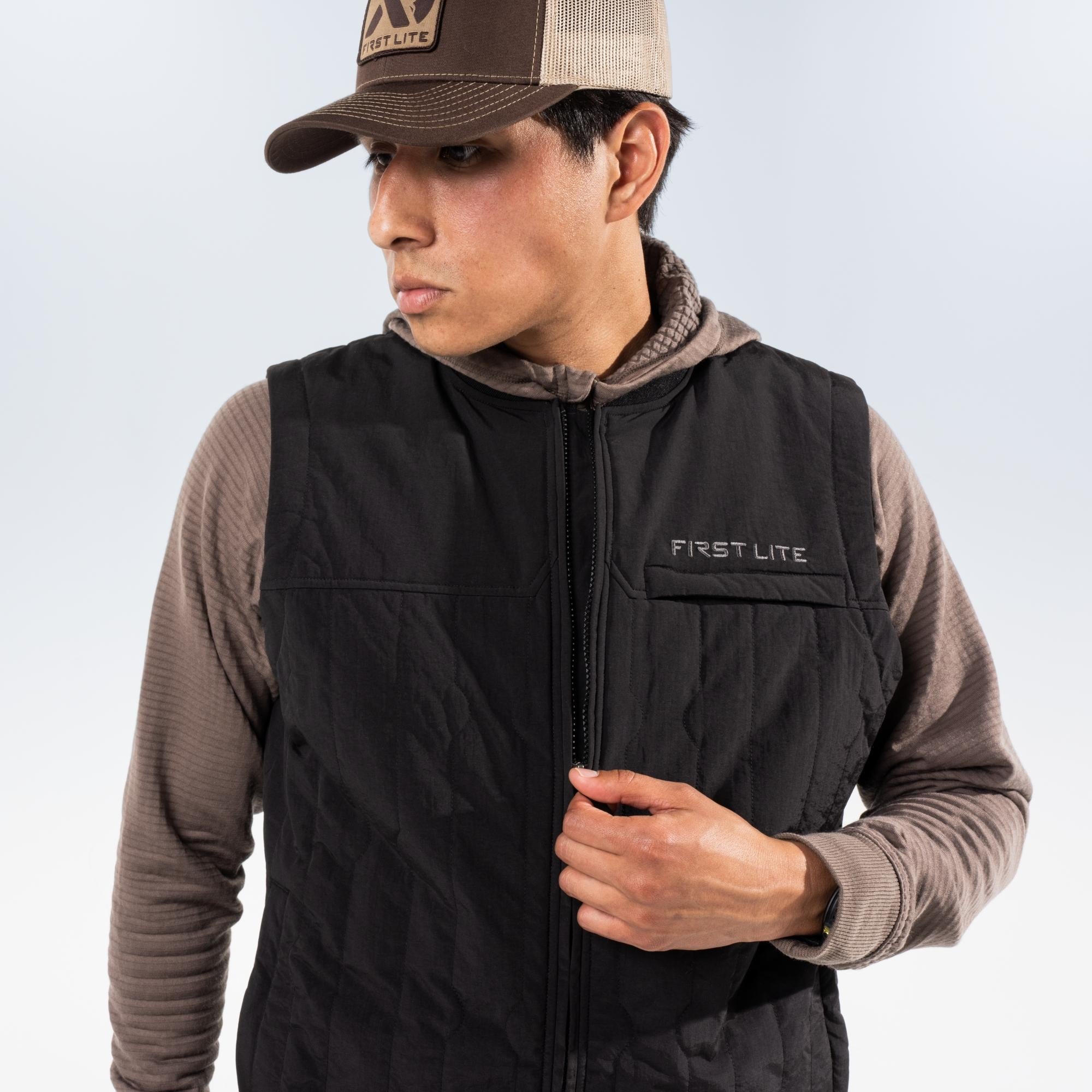Man wearing a brown long-sleeve shirt under a black vest and a brOWn FIRST LITE cap, standing against a light gray studio background.