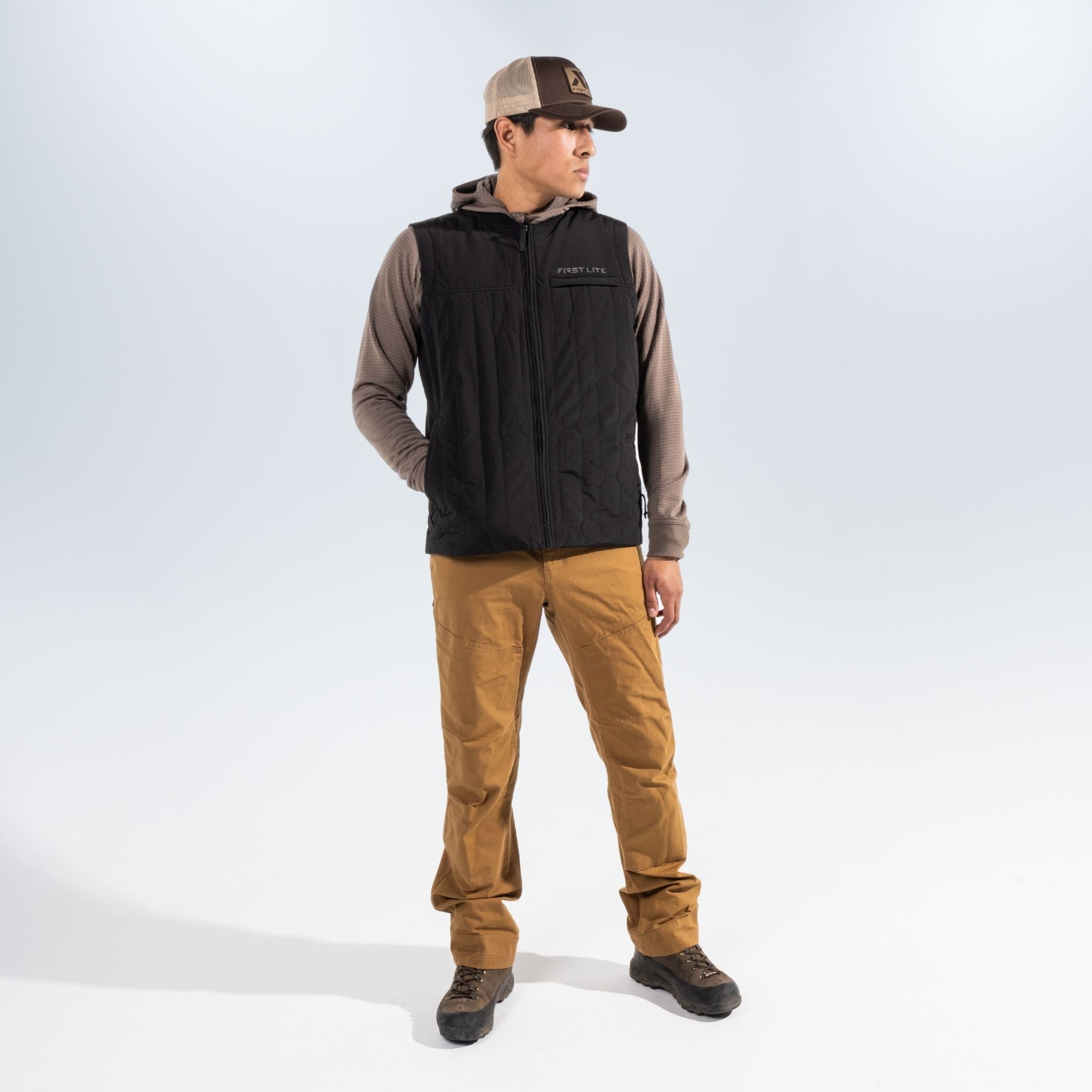 Man wearing a black vest over a brown hoodie, tan pants and brown boots standing against a white studio background.