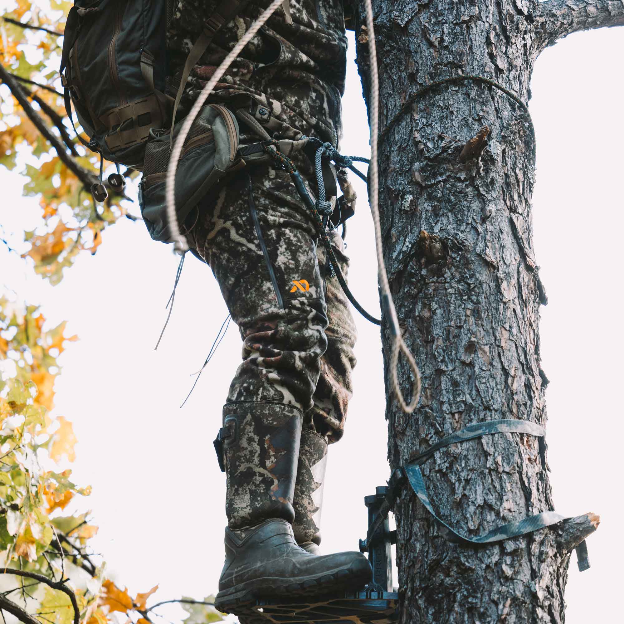 Man standing on a base strapped to a tree wearing FIRST LITE camo pants.