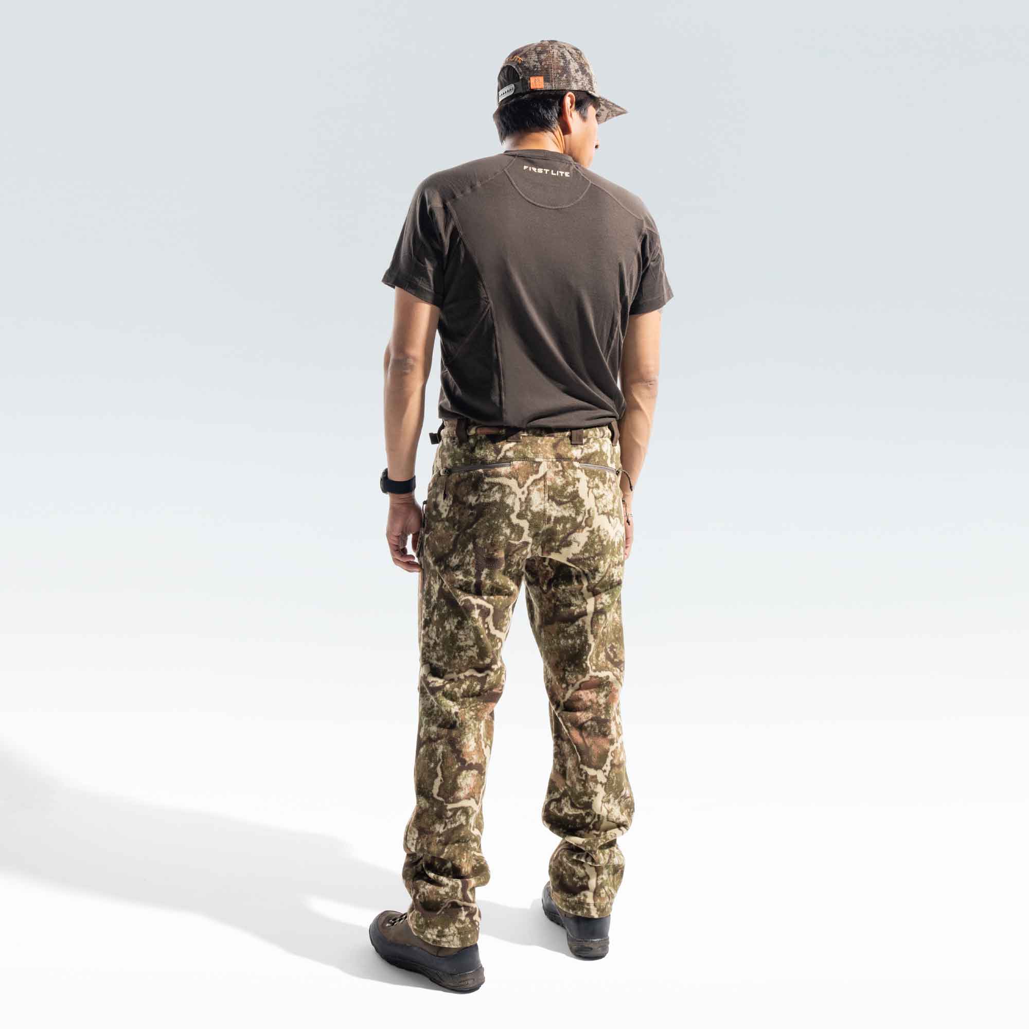Behind shot of a man wearing a FIRST LITE brown shirt, camo cap and camo pants. 