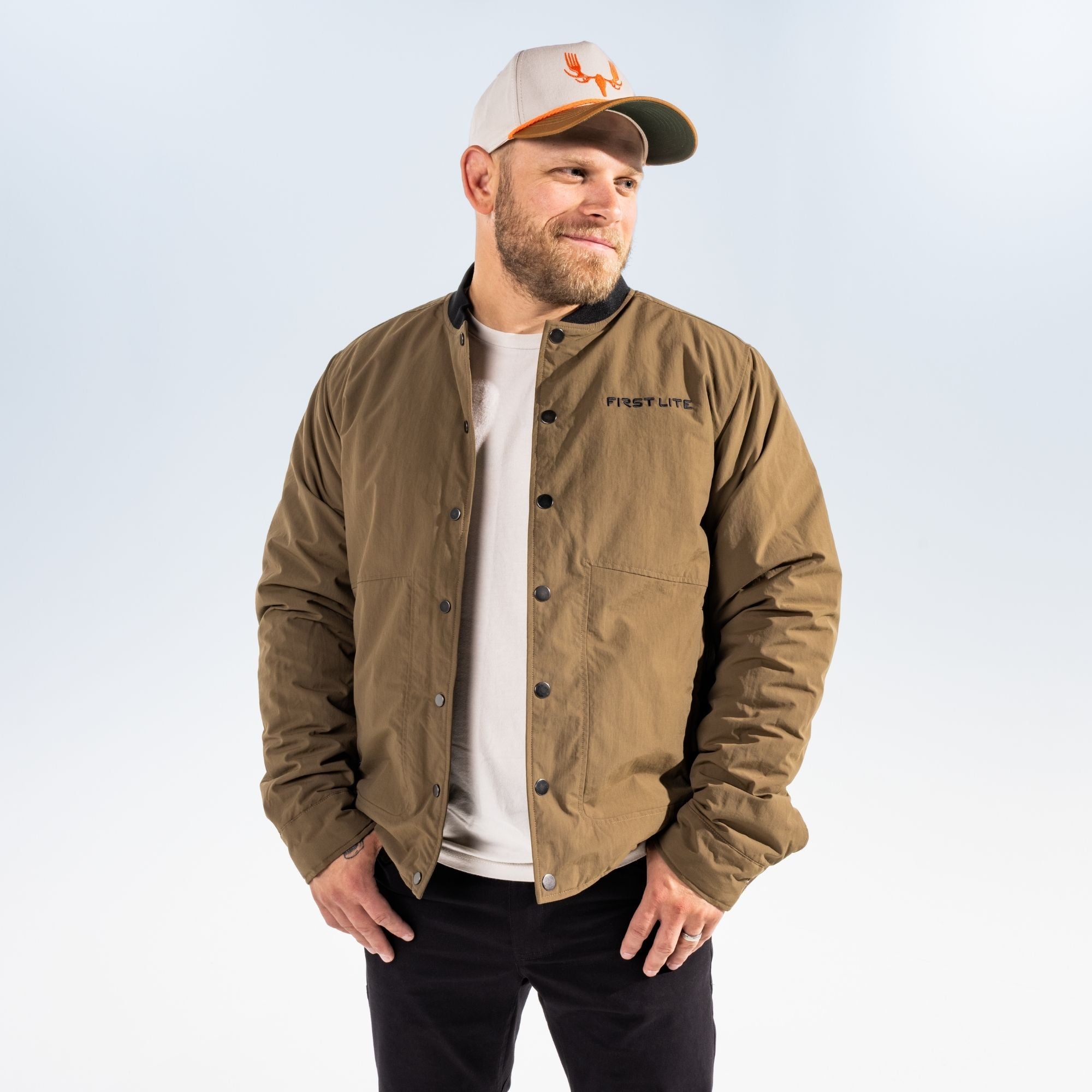 Man wearing an akern chore coat, a hat with MEATEATER logo in orange and black pants against a light gray studio background
