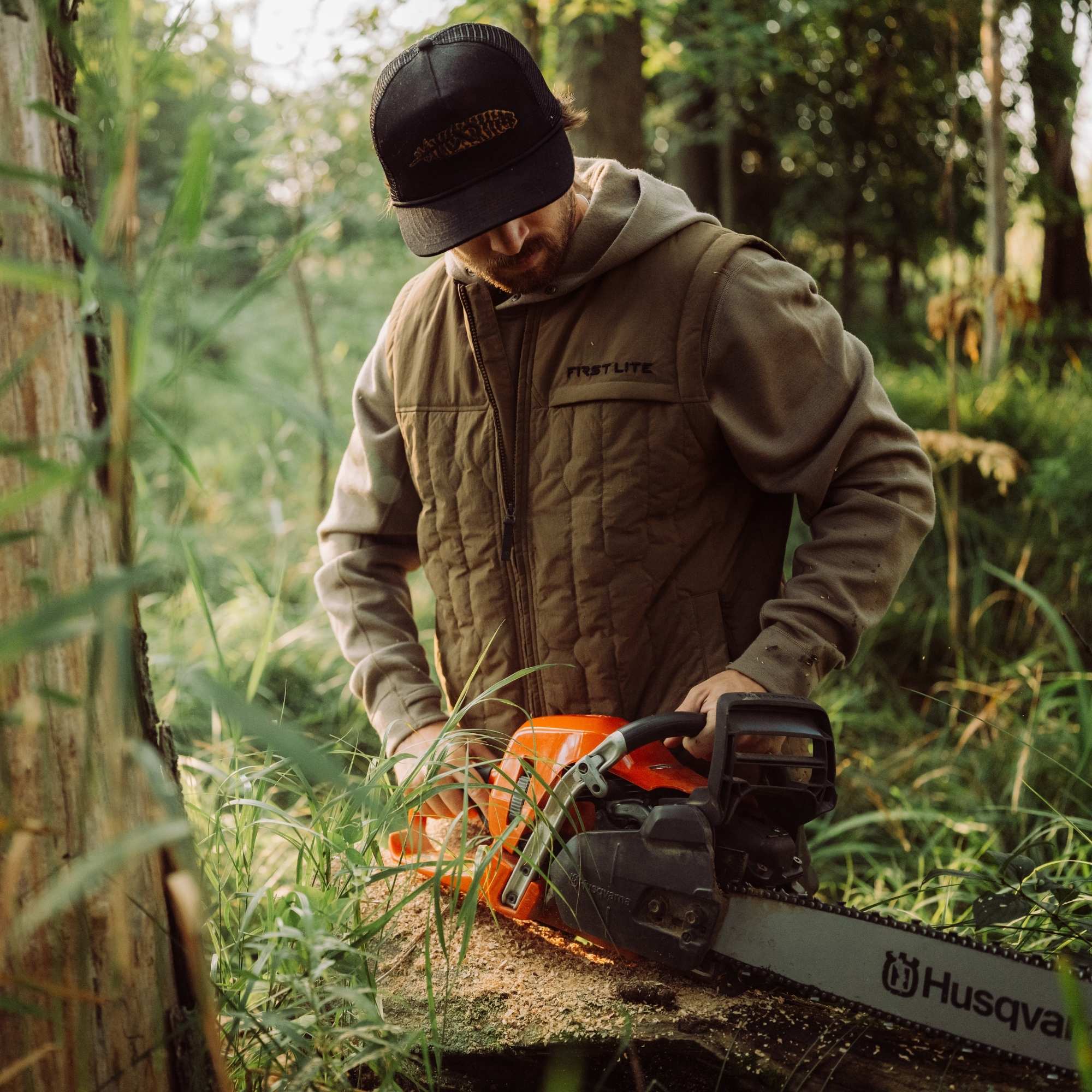 Man wearing an akern vest over a gray hoodie and a black cap holding a chainsaw in the forest.