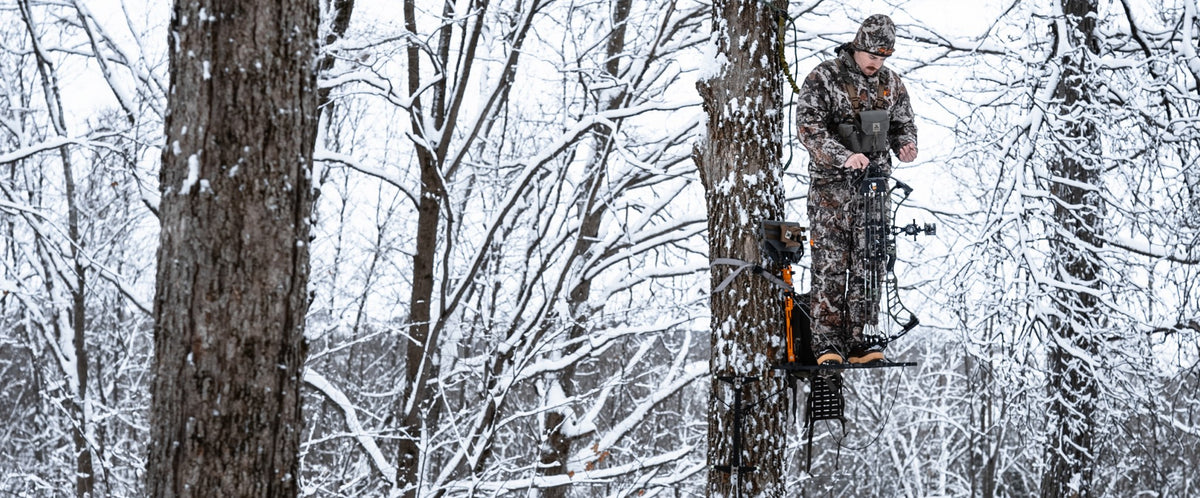 A person in camouflage stands on a tree platform in a snowy forest, holding a compound bow. Snow-covered trees fill the background in a winter outdoor setting.