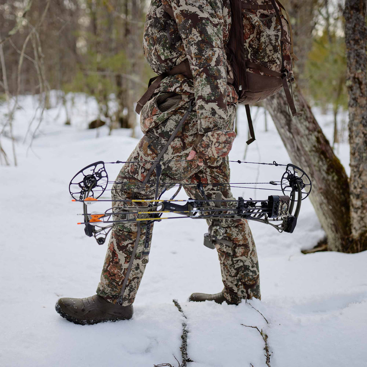 Man walking on a snowy forest carring bow and arrows wearing camo back pack, jacket and pants.