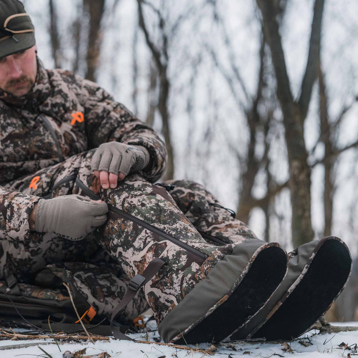 A man in camouflage winter hunting clothing sits in the snow outdoors, adjusting a pair of insulated hunting boots with straps and gloves.