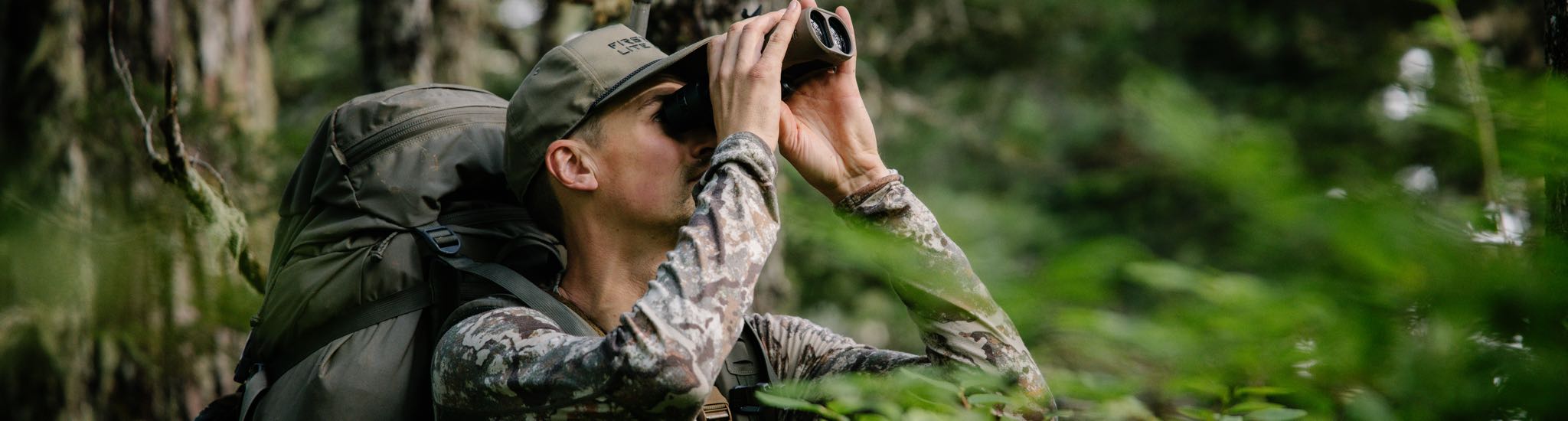 Person in camo shirt, green cap, using brown binoculars, with large green backpack in forest.